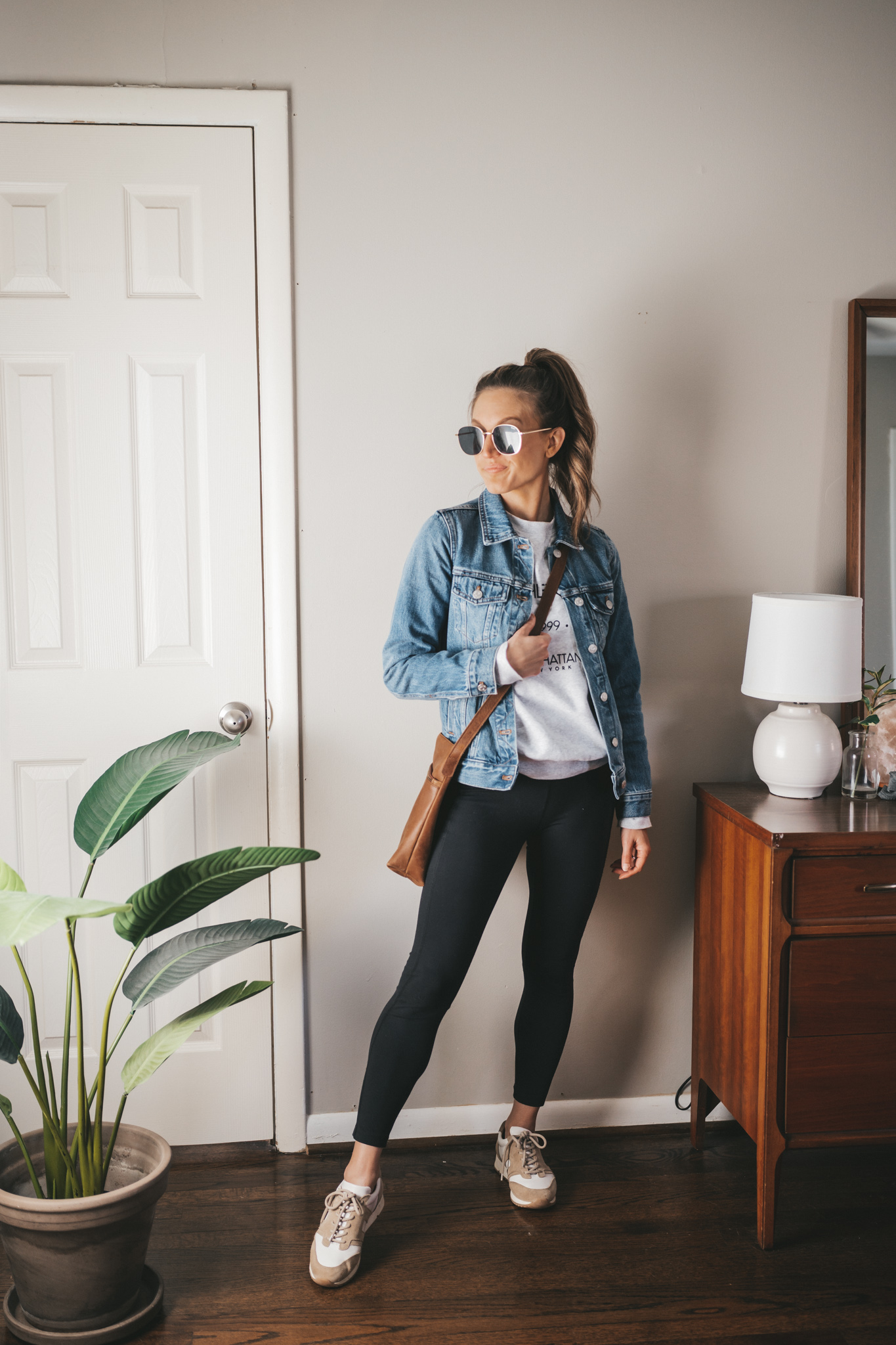 ashley wearing a gray graphic sweatshirt, denim jacket, black leggings, white and tan leather sneakers, and a brown crossbody bag with oversized gold frame sunglasses