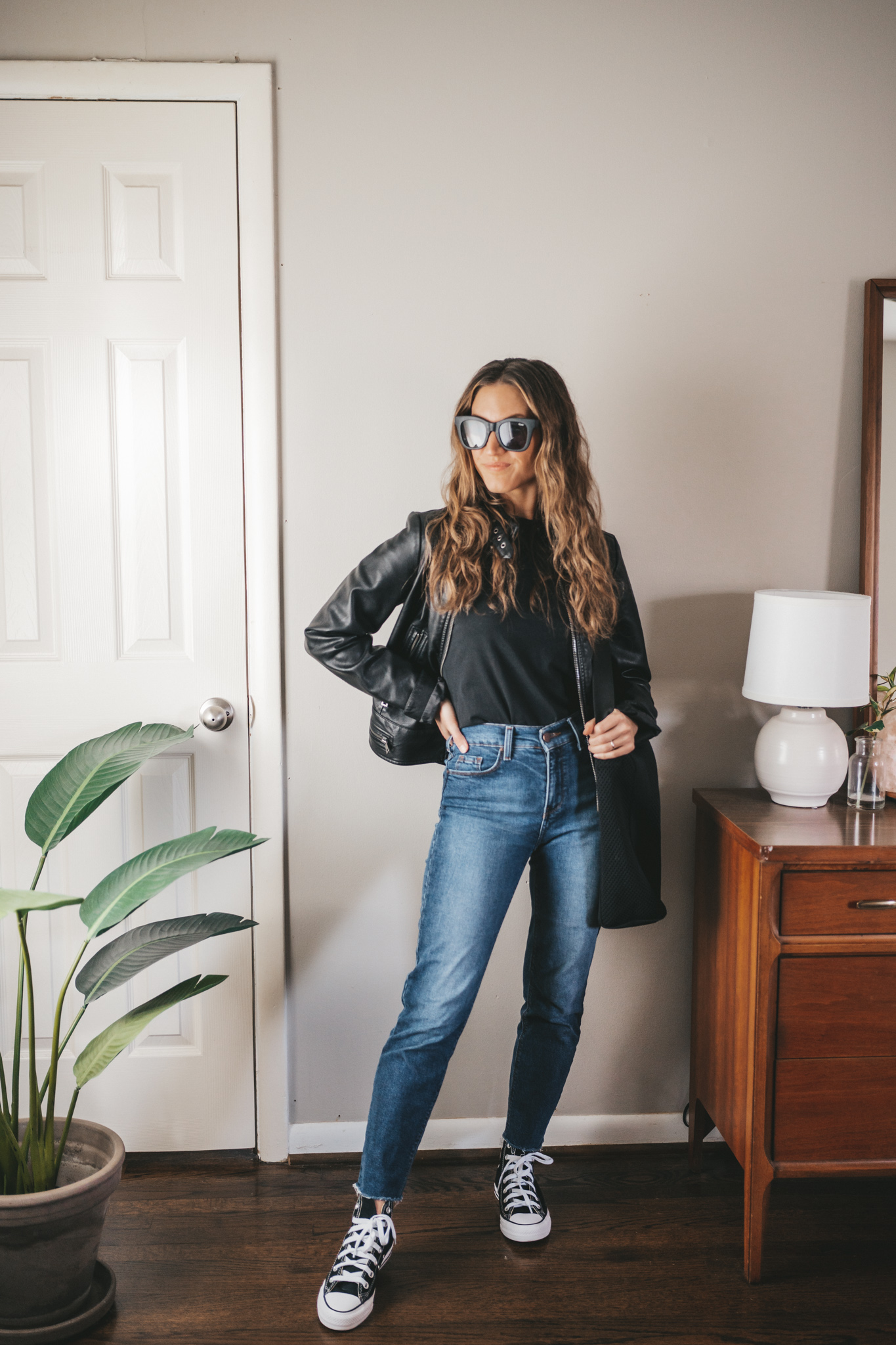 spring outfit idea with a black tee, faux leather moto jacket, straight denim, and black high top Converse, a black tote bag, and black square frame sunglasses