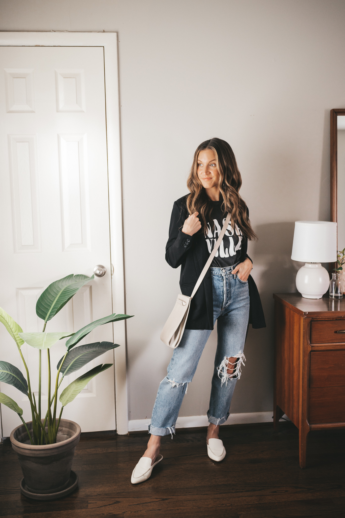 spring outfit idea with a black graphic tee, black boyfriend blazer, distressed denim, white mules, and a gray crossbody bag