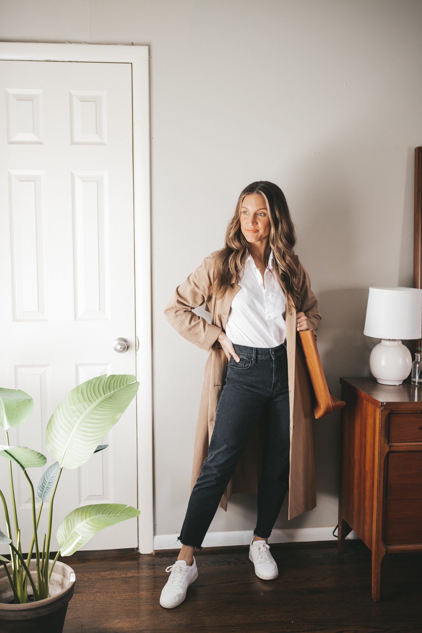 ashley wearing a white button down, tan trench coat, black straight denim, and white sneakers with a cognac crossbody bag