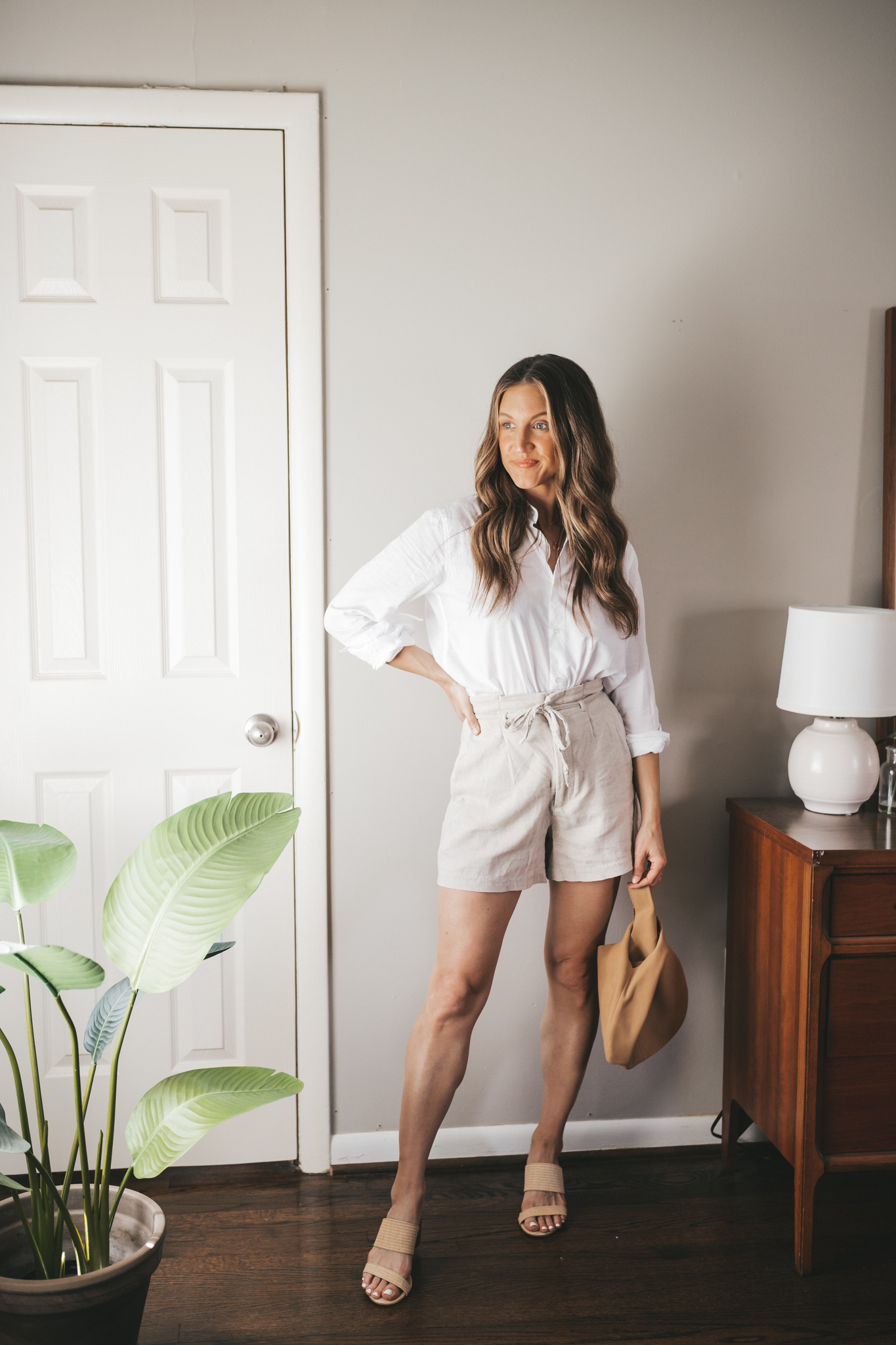 ashley wearing a white button down with tan linen shorts, heeled straw two strap sandals, and a tan leather clutch bag