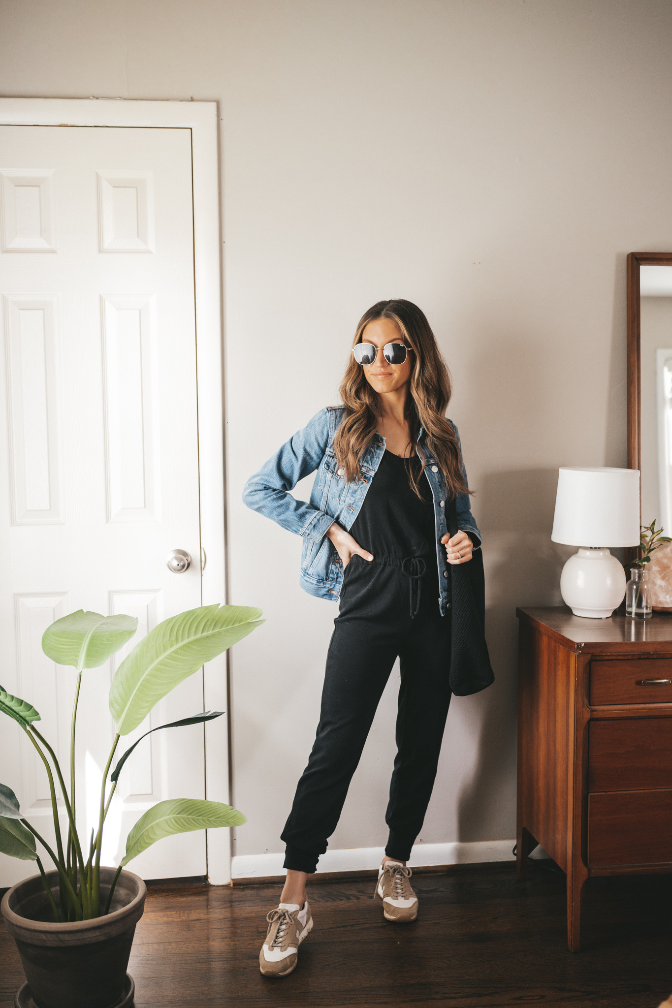 spring outfit idea with a black jumpsuit, denim jacket, white and tan leather sneakers, a black tote bag, and oversized gold frame sunglasses