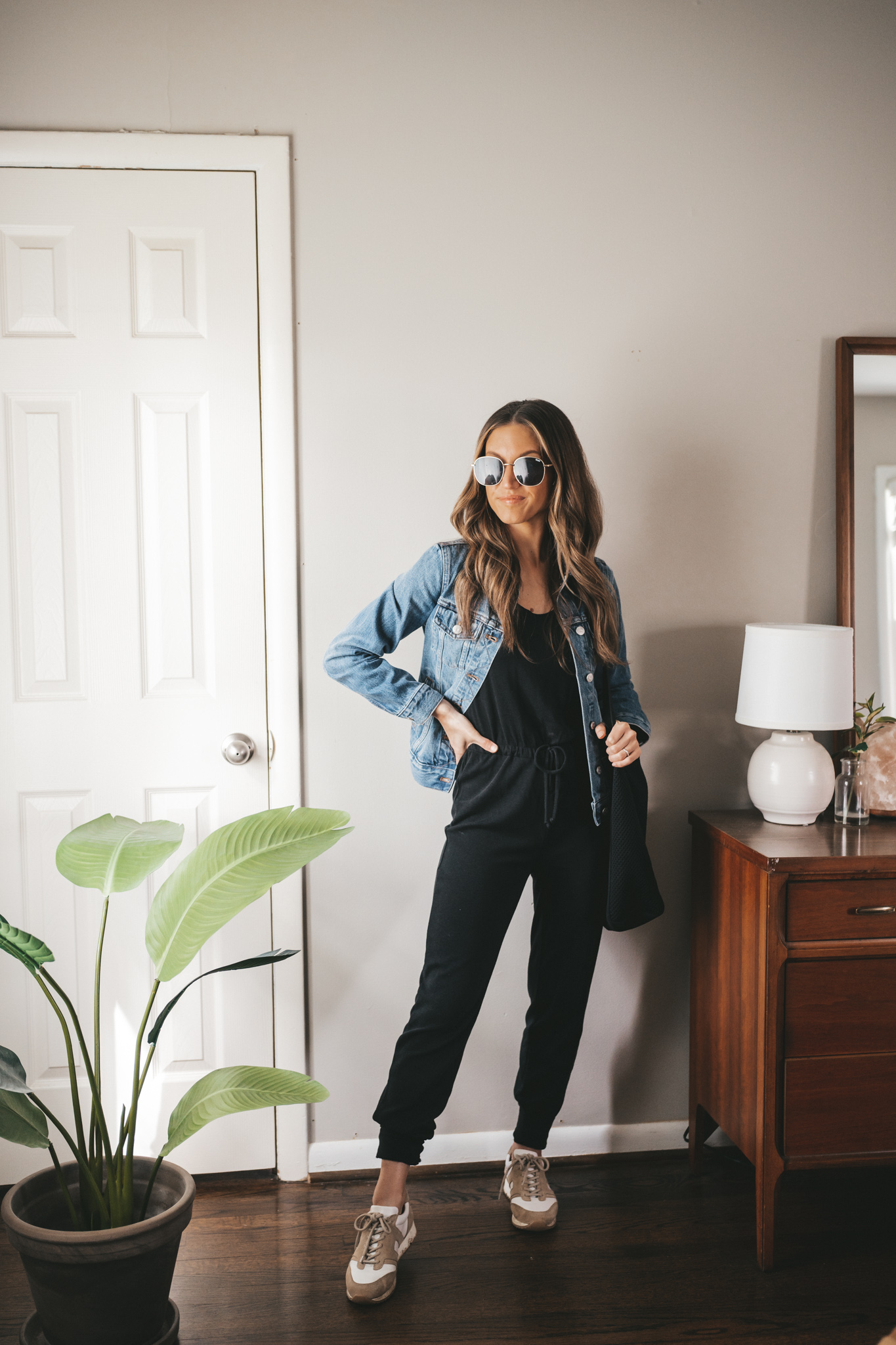 spring outfit idea with a black jumpsuit, denim jacket, white and tan leather sneakers, a black tote bag, and oversized gold frame sunglasses