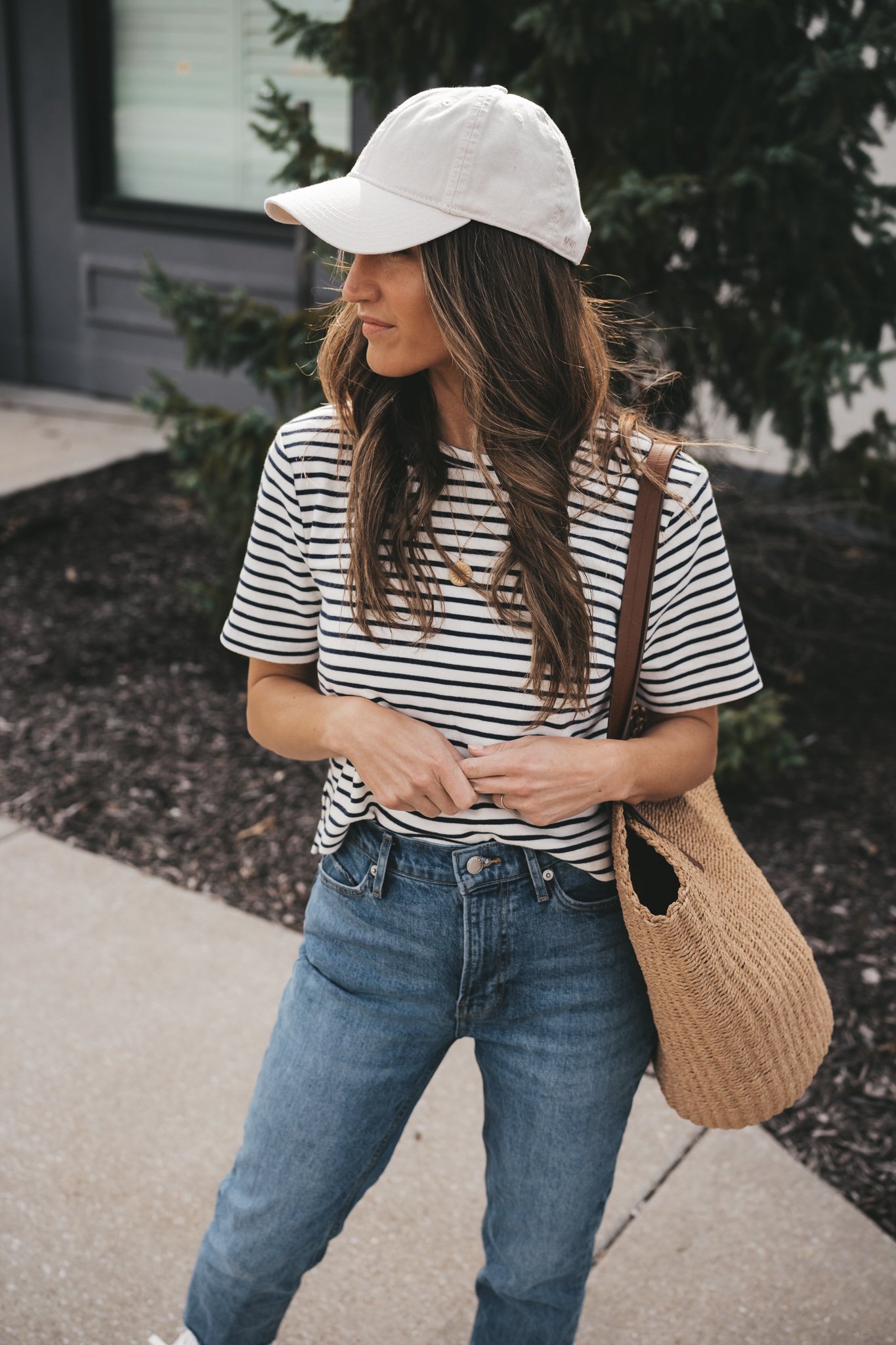 ashley standing on a sidewalk in front of a gray building wearing a white and navy striped top, denim, white sneakers, a straw bag, and ivory baseball cap