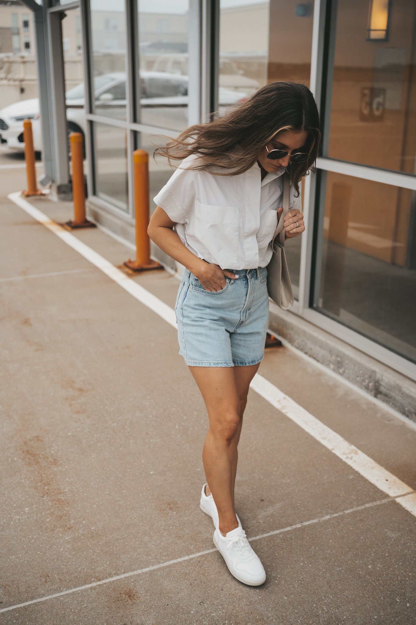 ashley wearing a light gray button down top tucked into the Everlane A-Line Denim shorts with white sneakers and oversized sunglasses standing in front of glass windows