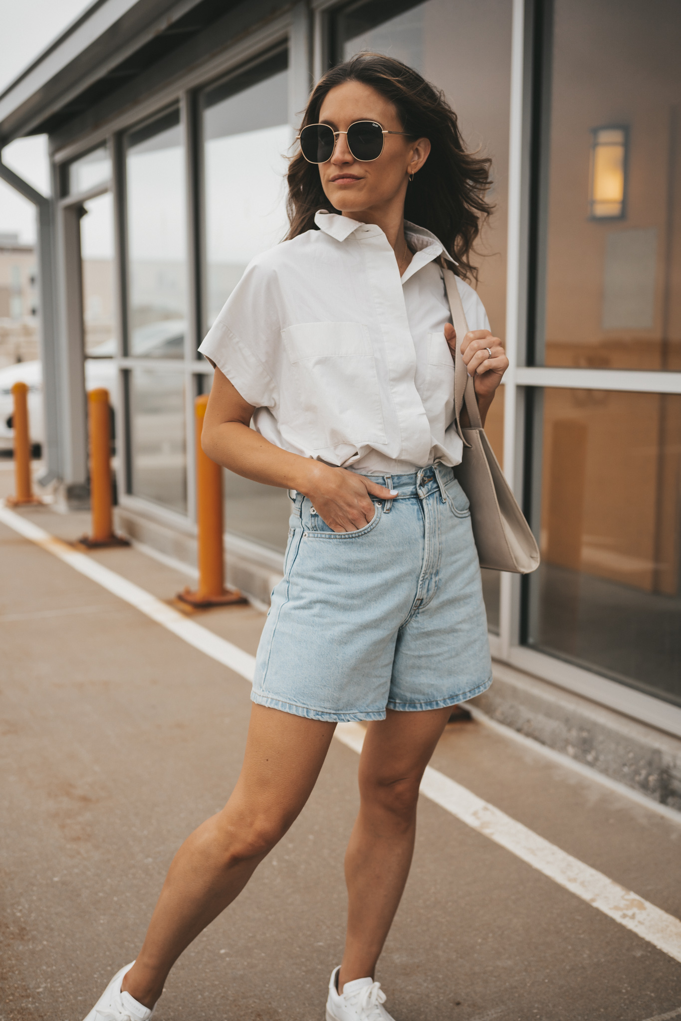 ashley wearing a light gray button down top tucked into the Everlane A-Line Denim shorts with white sneakers and oversized sunglasses standing in front of glass windows