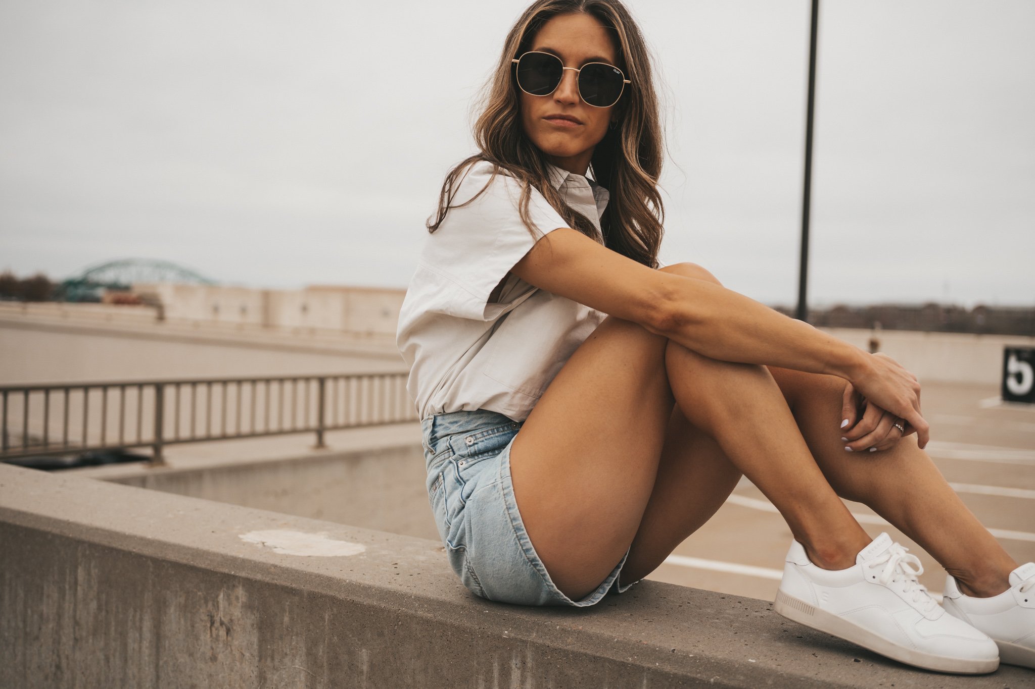 ashley wearing a light gray button down top tucked into the Everlane A-Line Denim shorts with white sneakers and oversized sunglasses sitting on a concrete wall