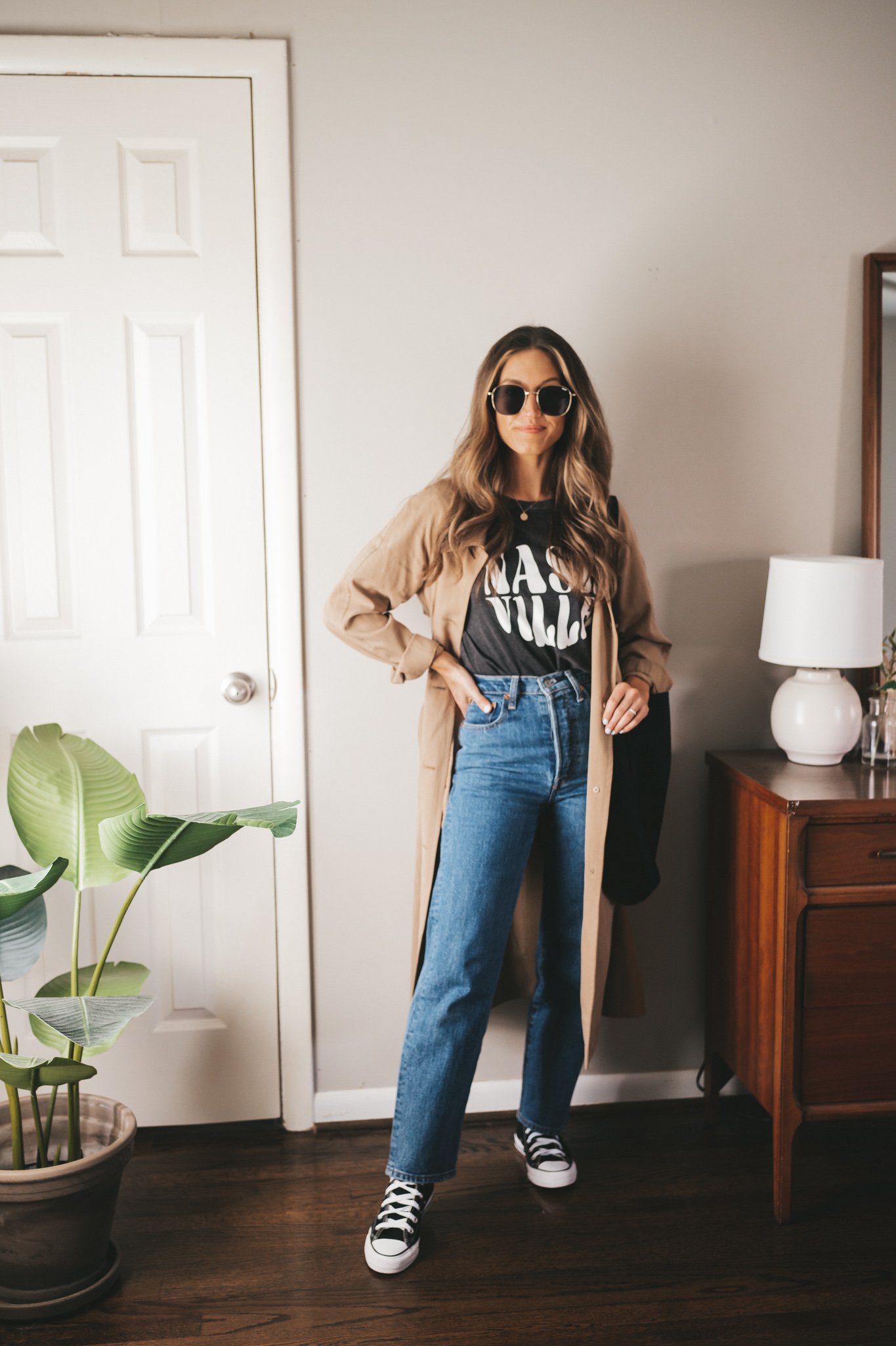 ashley wearing a black graphic tee, tan trench coat, full length mid-wash denim, and black high top Converse