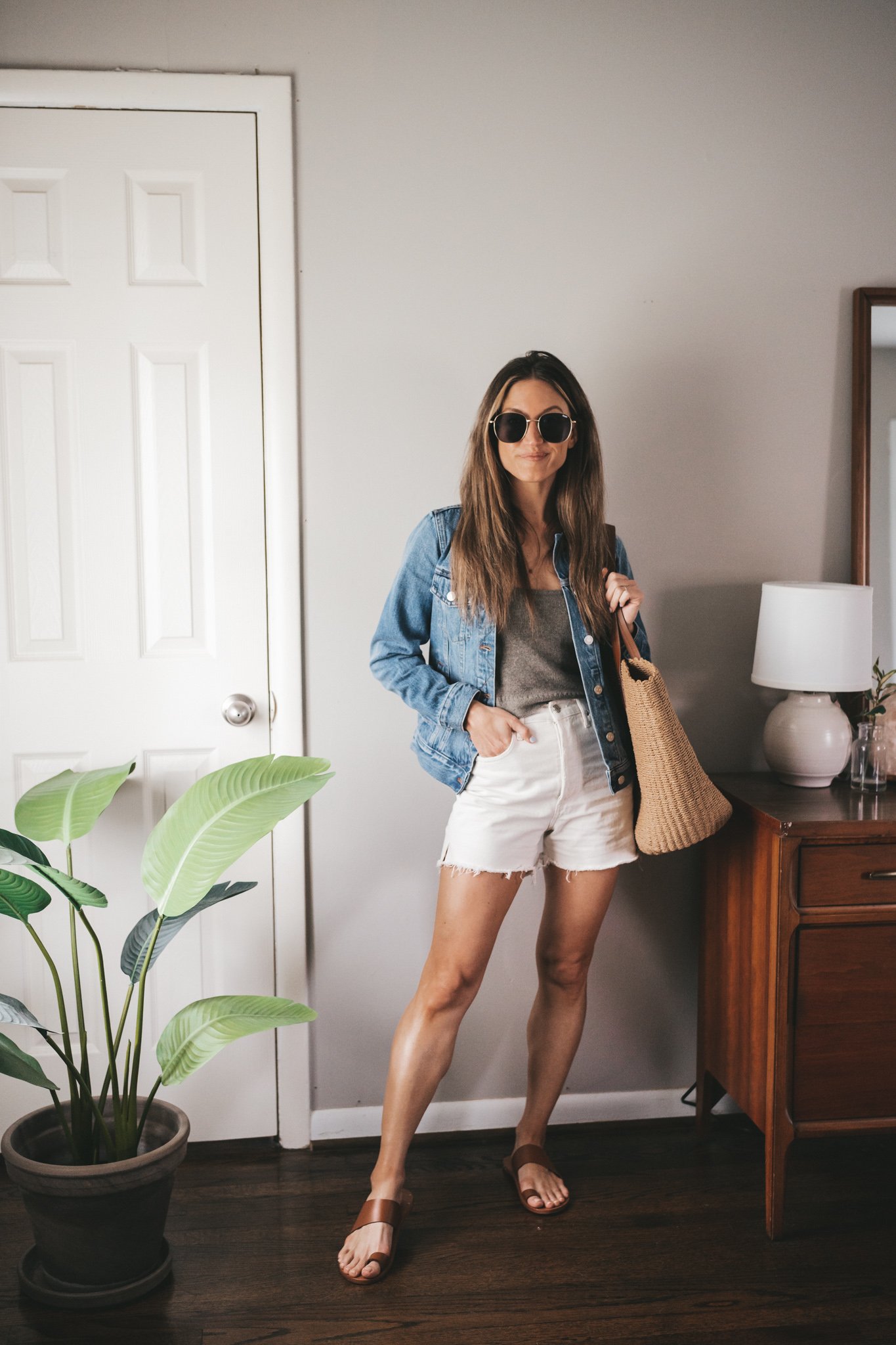 outfit for spring with a taupe sweater tank, denim jacket, white denim shorts, and brown sandals with a straw bag