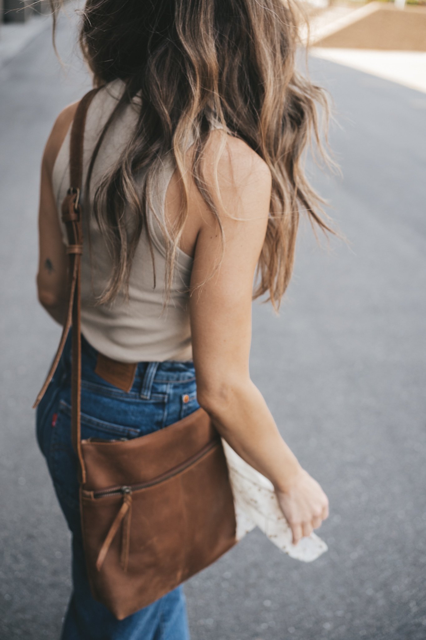 ashley wearing a tan tank, mid-wash denim, braided sandals and a brown crossbody bag with a white scarf tied around the strap