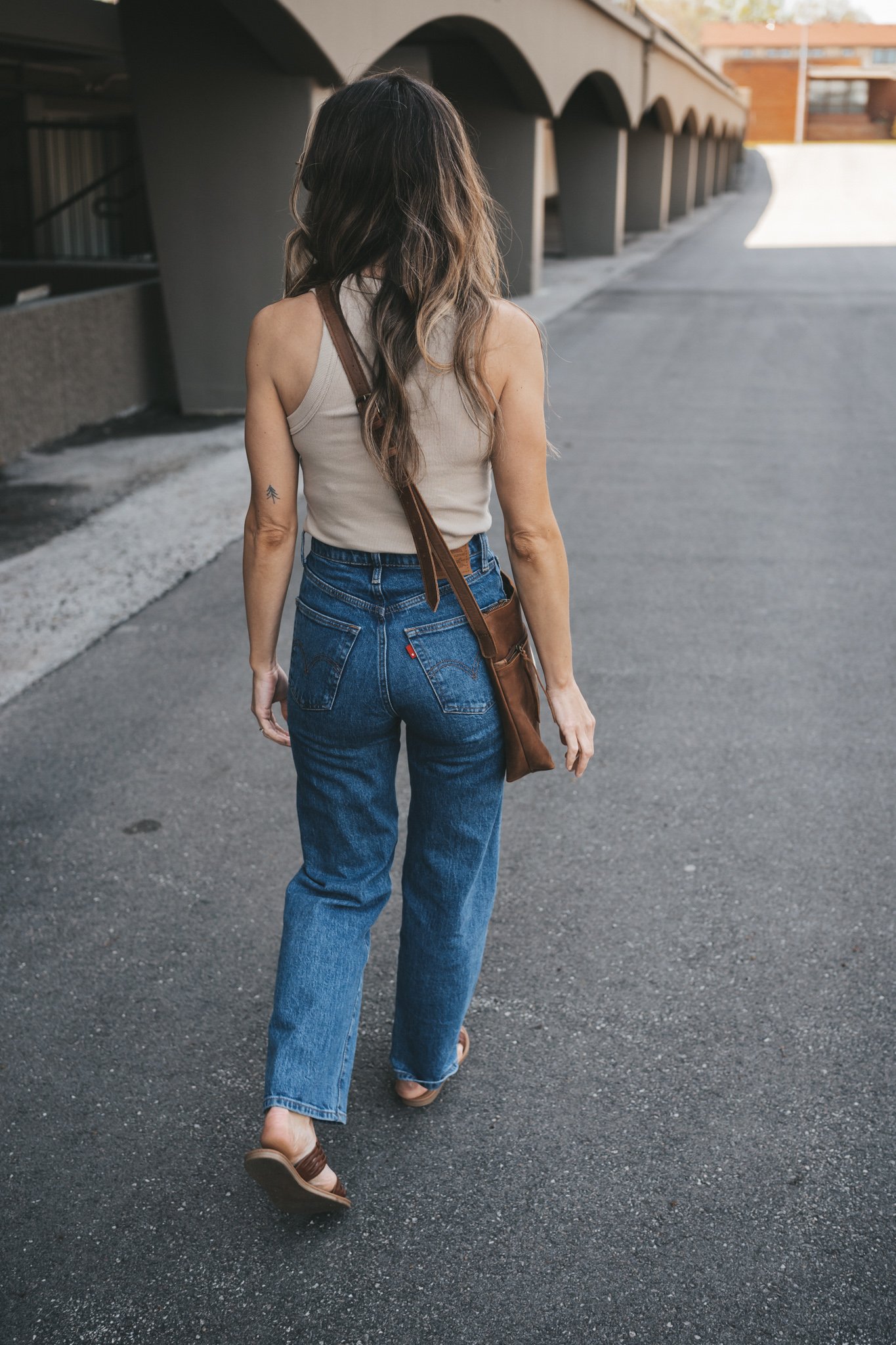 ashley wearing a tan tank, mid-wash denim, braided sandals and a brown crossbody bag with a white scarf tied around the strap