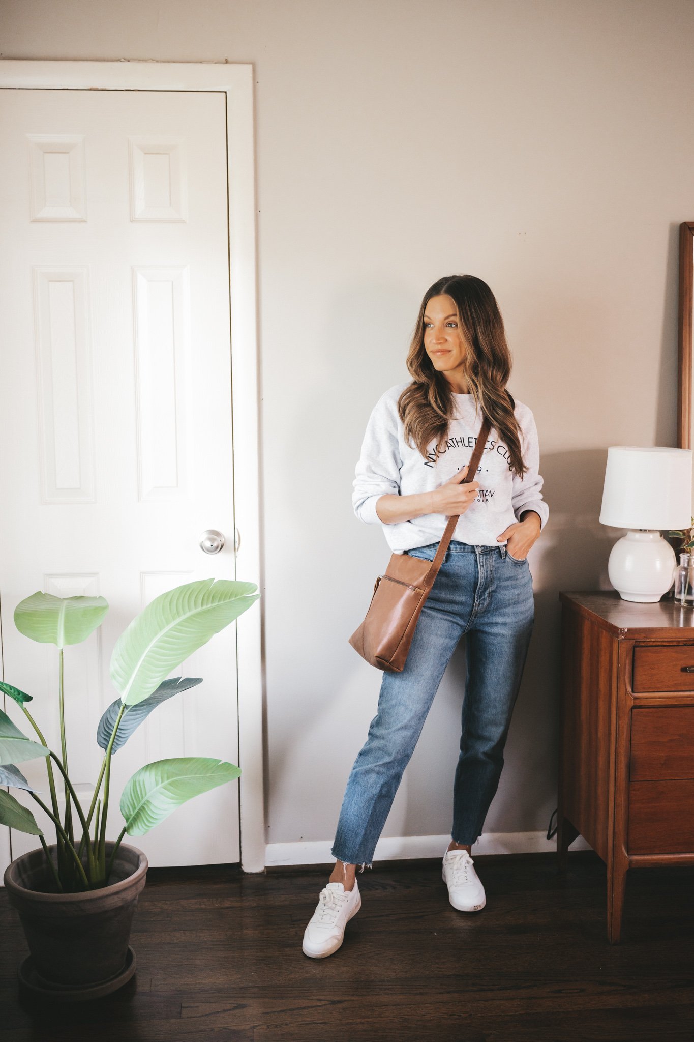 ashley wearing a gray graphic sweatshirt, mid-wash straight denim, and white sneakers with a brown leather crossbody bag