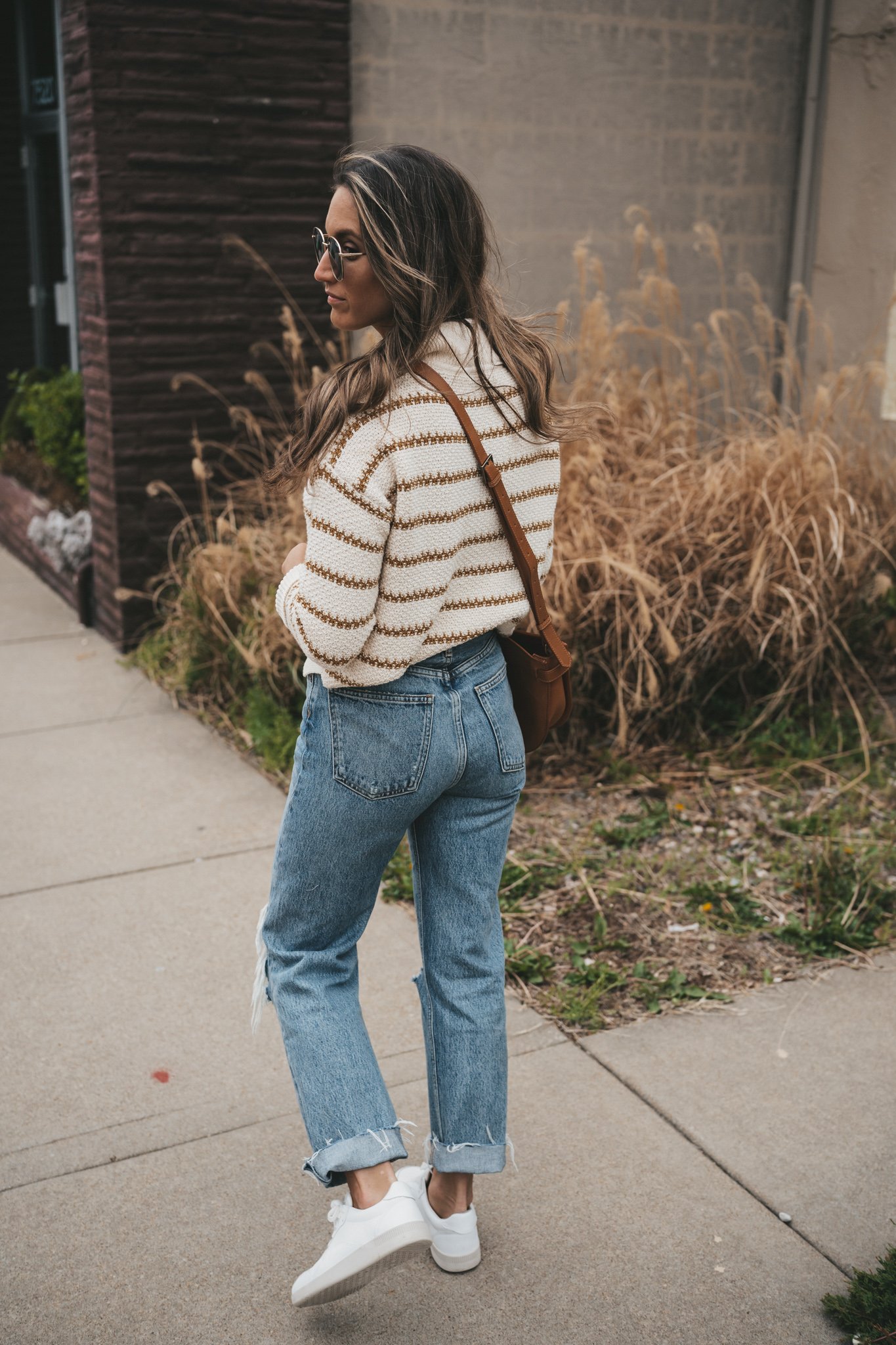 ashley wearing a striped sweater, distressed denim, white sneakers, and a cognac crossbody bag standing in front of a shop with plants