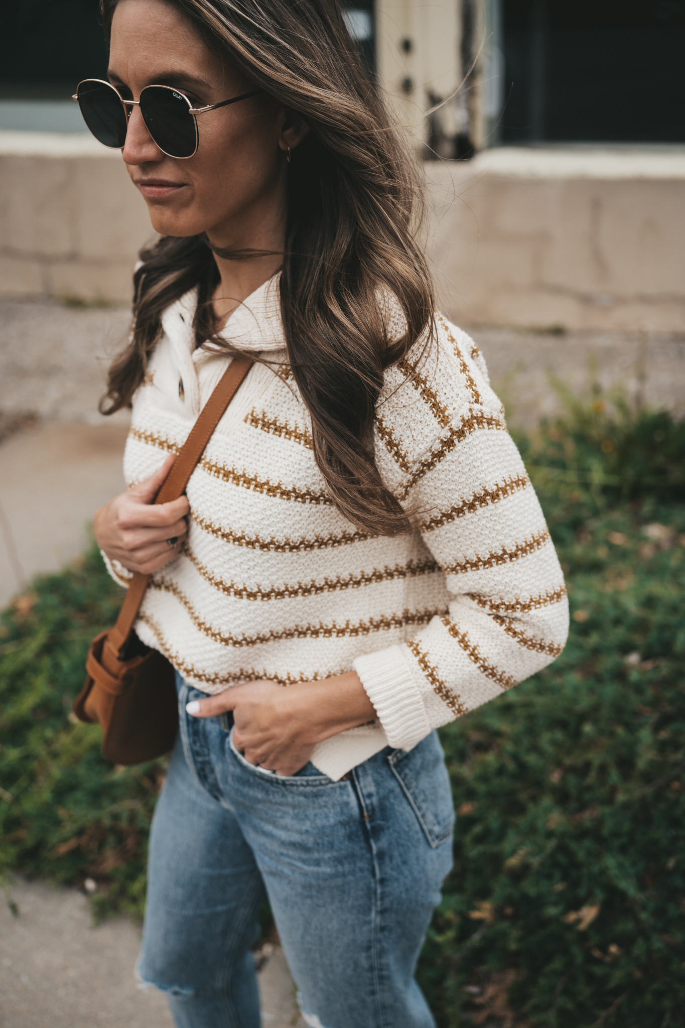 ashley wearing a striped sweater, distressed denim, white sneakers, and a cognac crossbody bag standing in front of a shop with plants