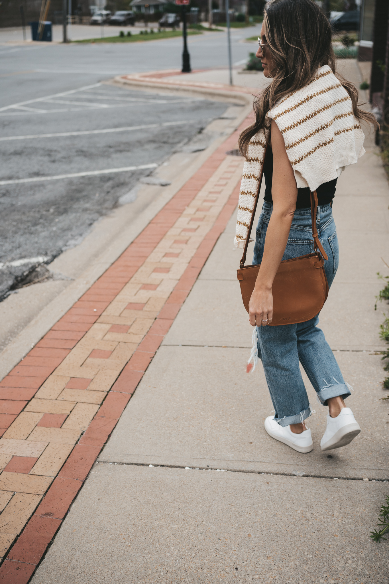 ashley wearing a striped sweater, distressed denim, white sneakers, and a cognac crossbody bag standing in front of a shop with plants