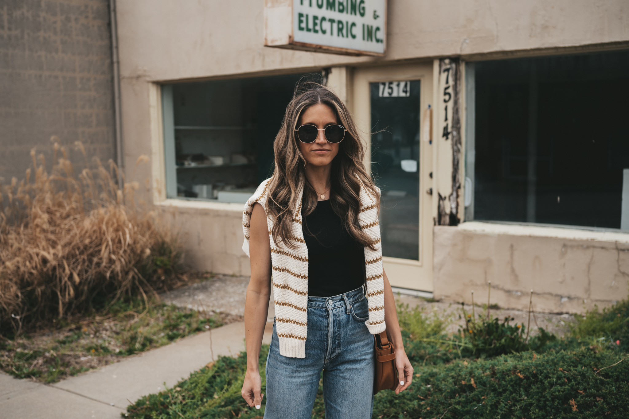 ashley wearing a striped sweater, distressed denim, white sneakers, and a cognac crossbody bag standing in front of a shop with plants