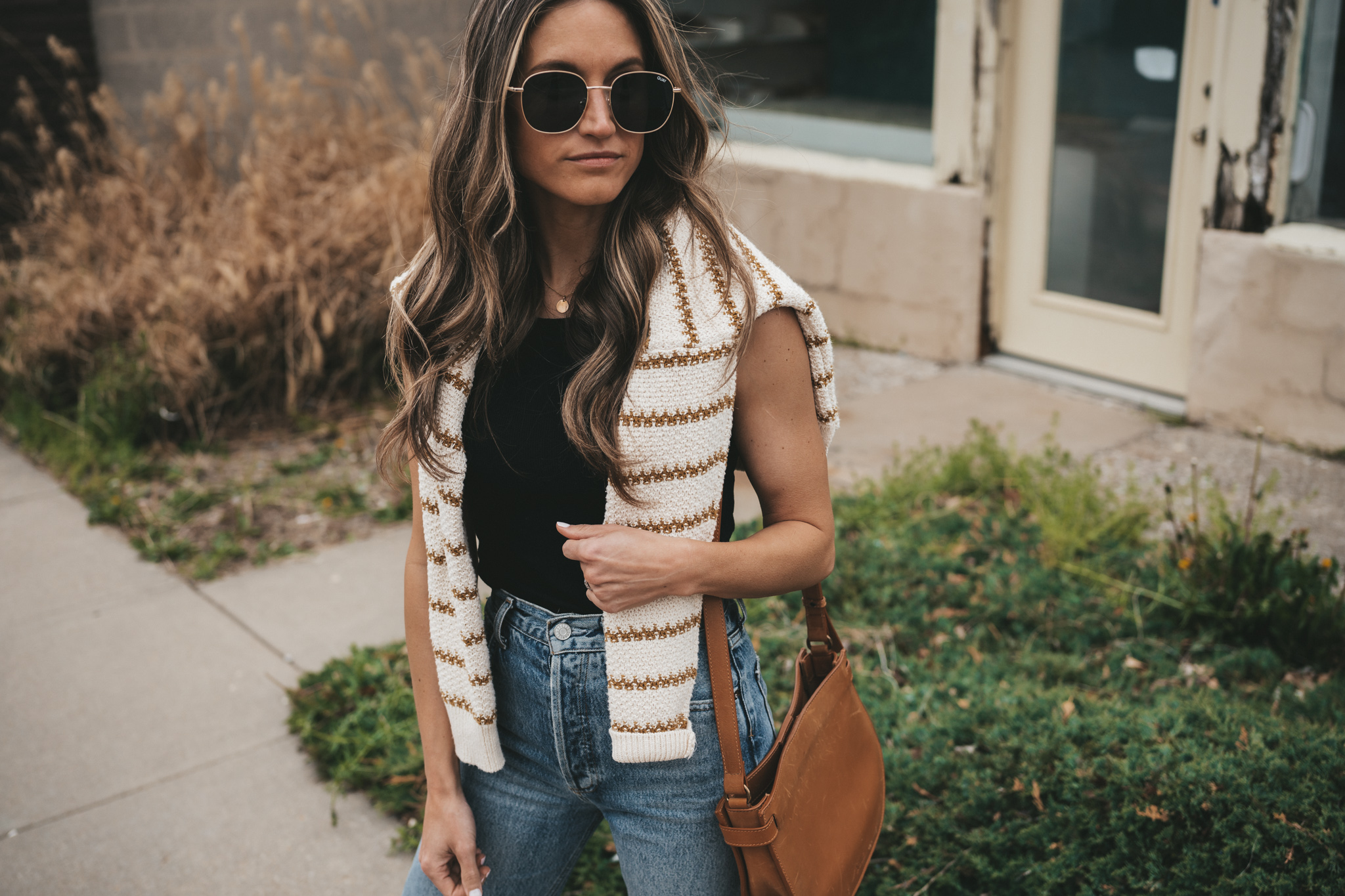 ashley wearing a striped sweater, distressed denim, white sneakers, and a cognac crossbody bag standing in front of a shop with plants