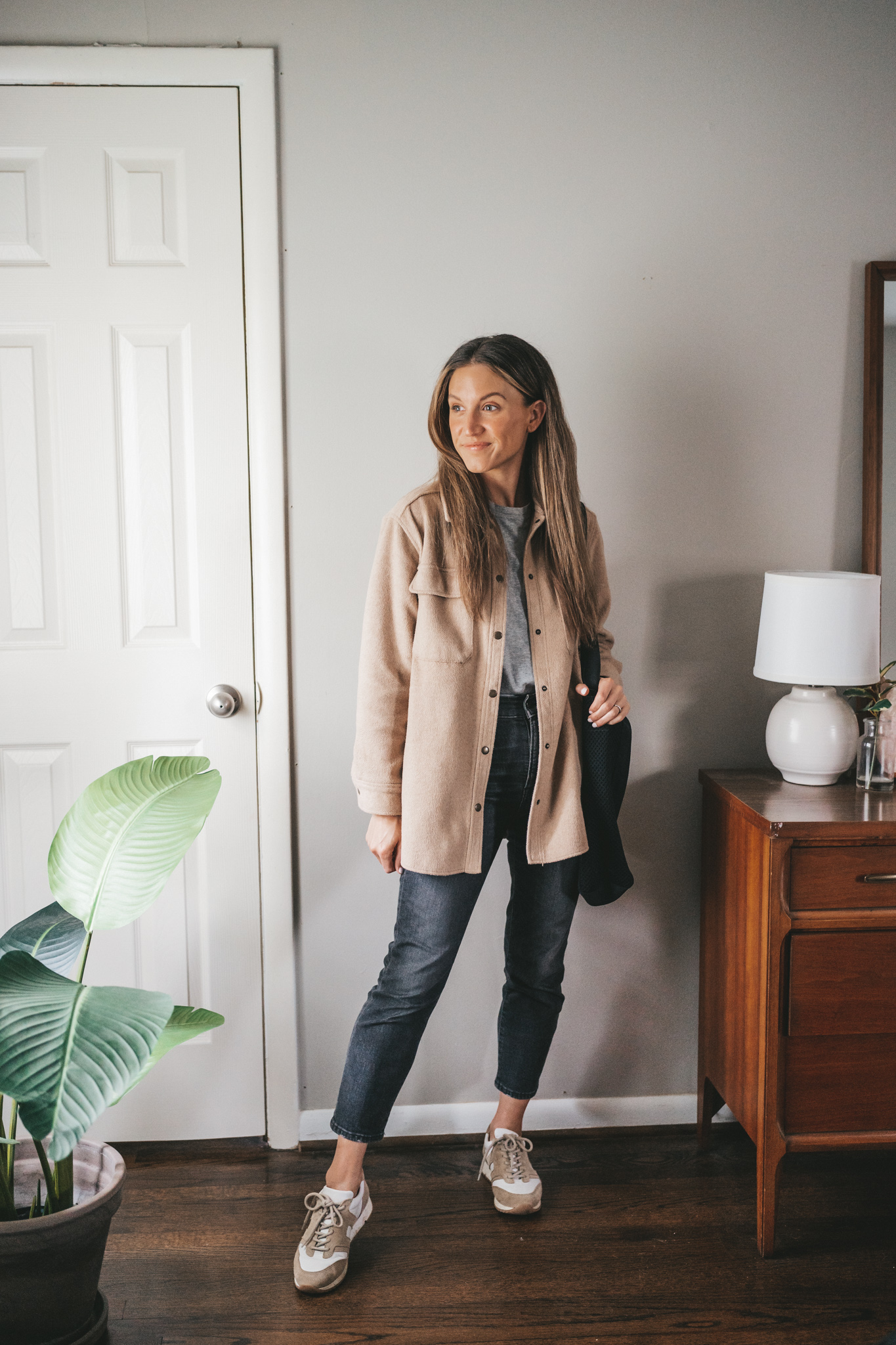 ashley wearing a gray t-shirt, washed black denim, tan shacket, and tan and white leather sneakers with a black tote bag