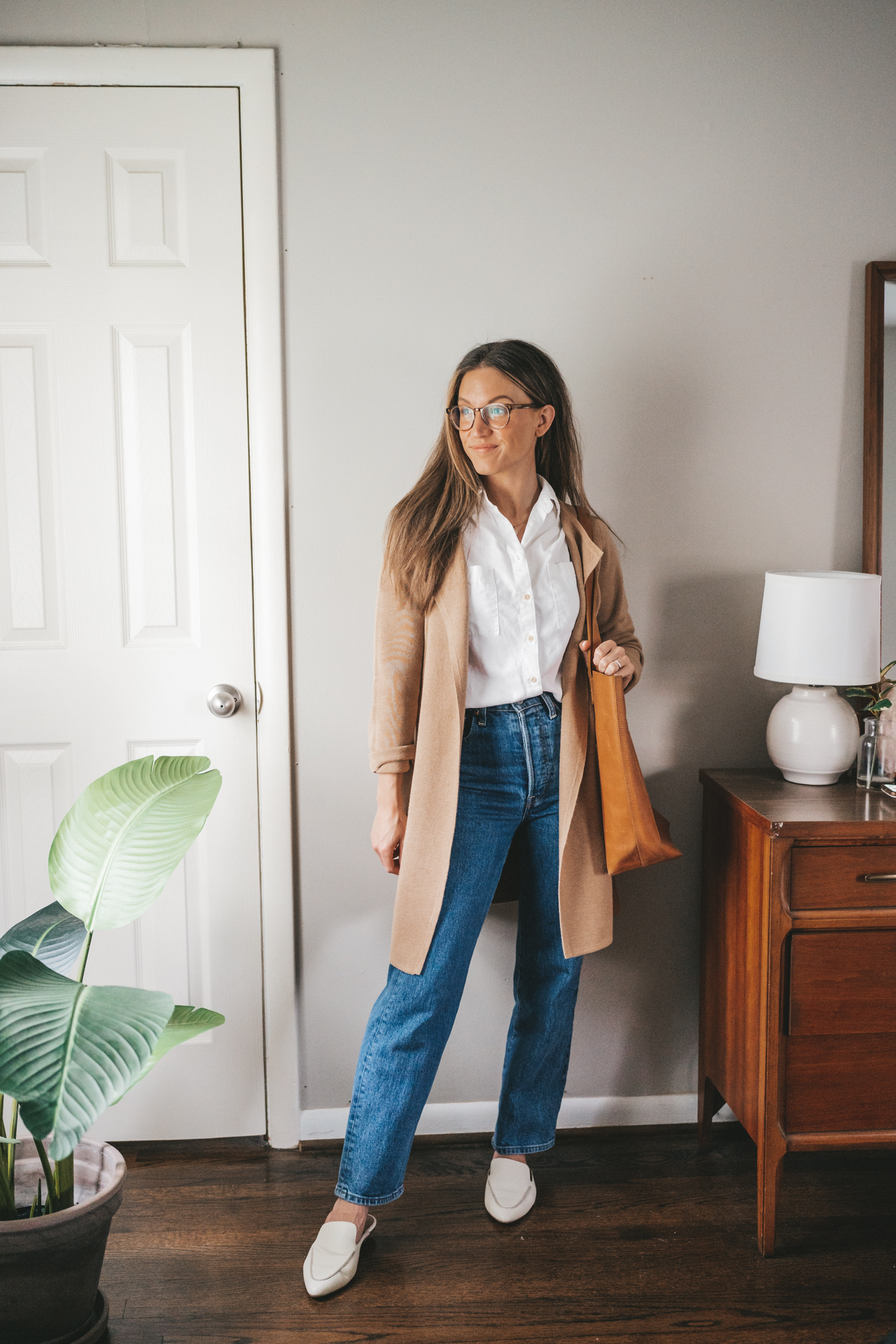ashley wearing a white button down, tan long line sweater blazer, mid-wash denim, and white mules with a cognac tote bag