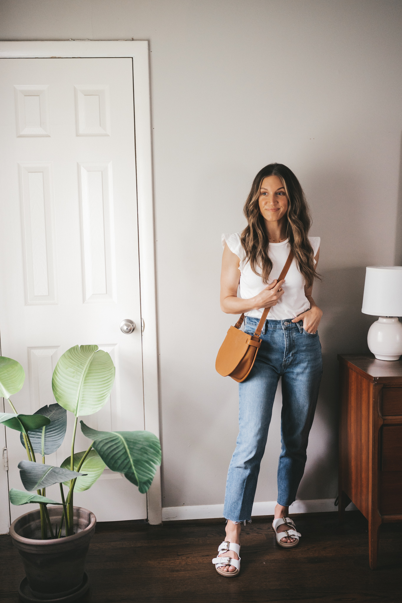 ashley wearing a white ruffle sleeve tank, mid-wash denim, white two strap sandals, and a cognac crossbody bag