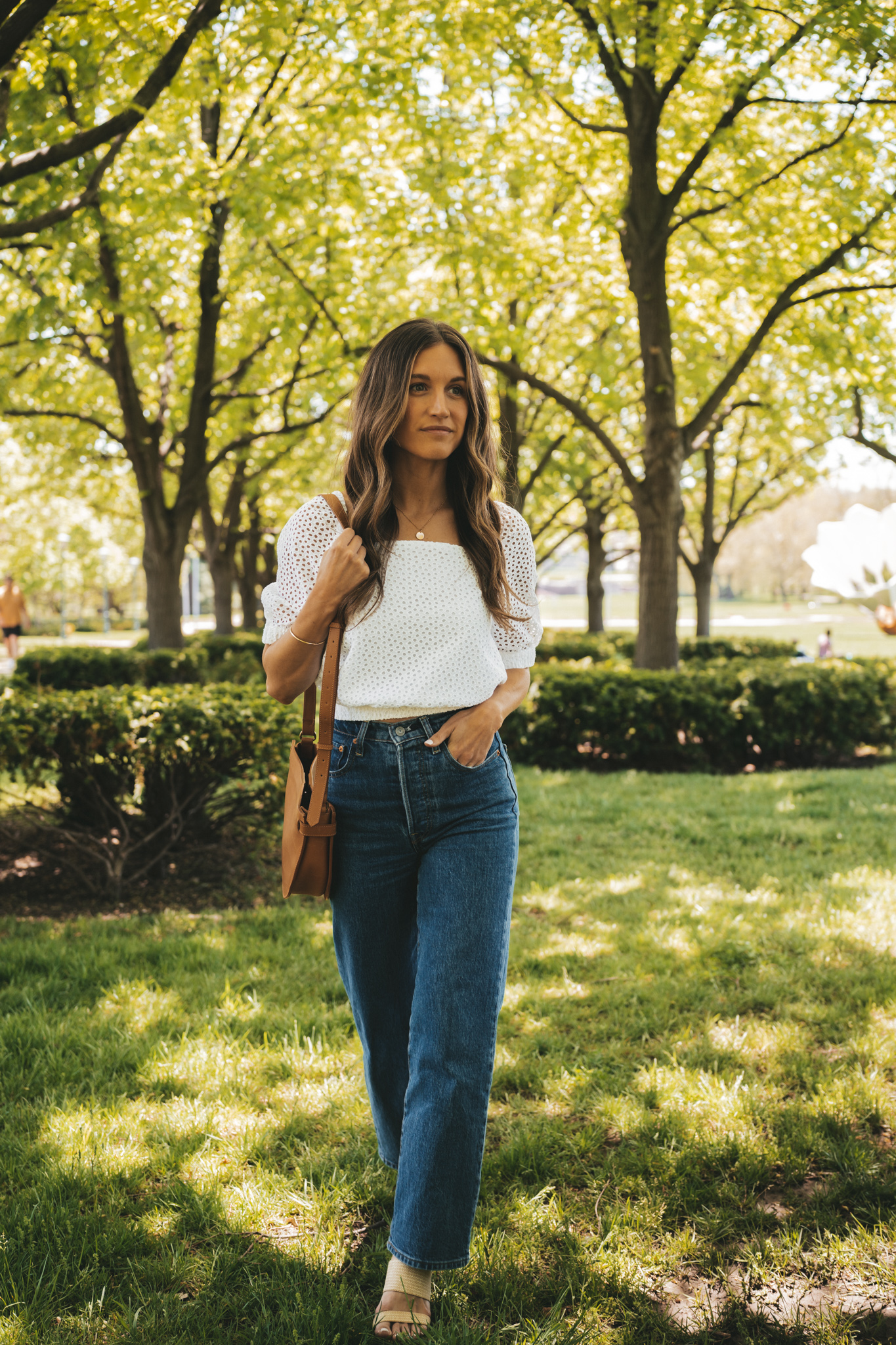 ashley wearing an eyelet top, mid-wash denim, woven heels, and a cognac crossbody bag in a park