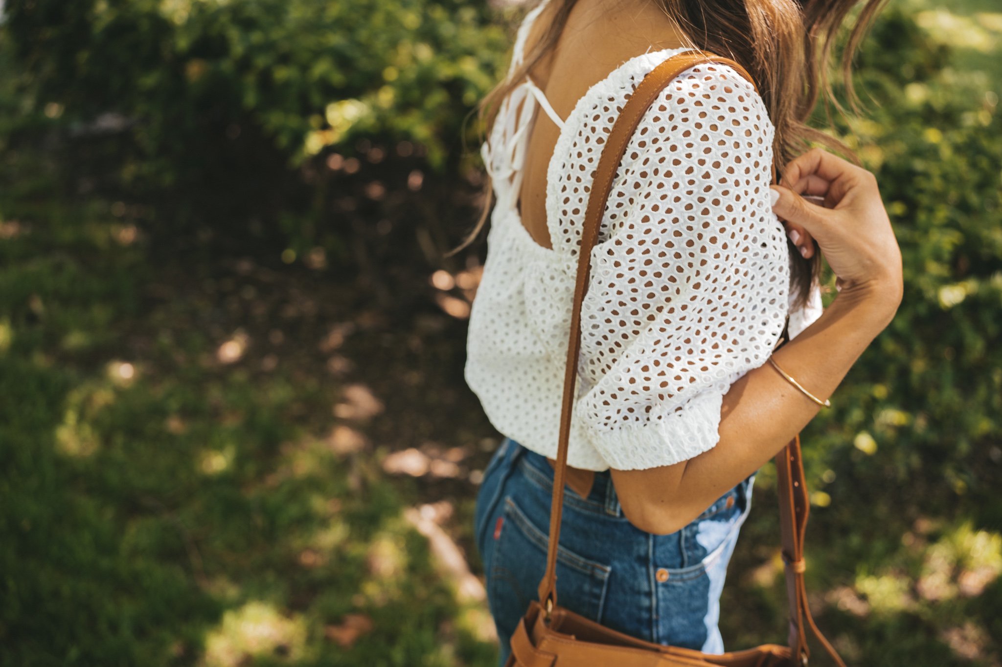 ashley wearing an eyelet top, mid-wash denim, woven heels, and a cognac crossbody bag in a park