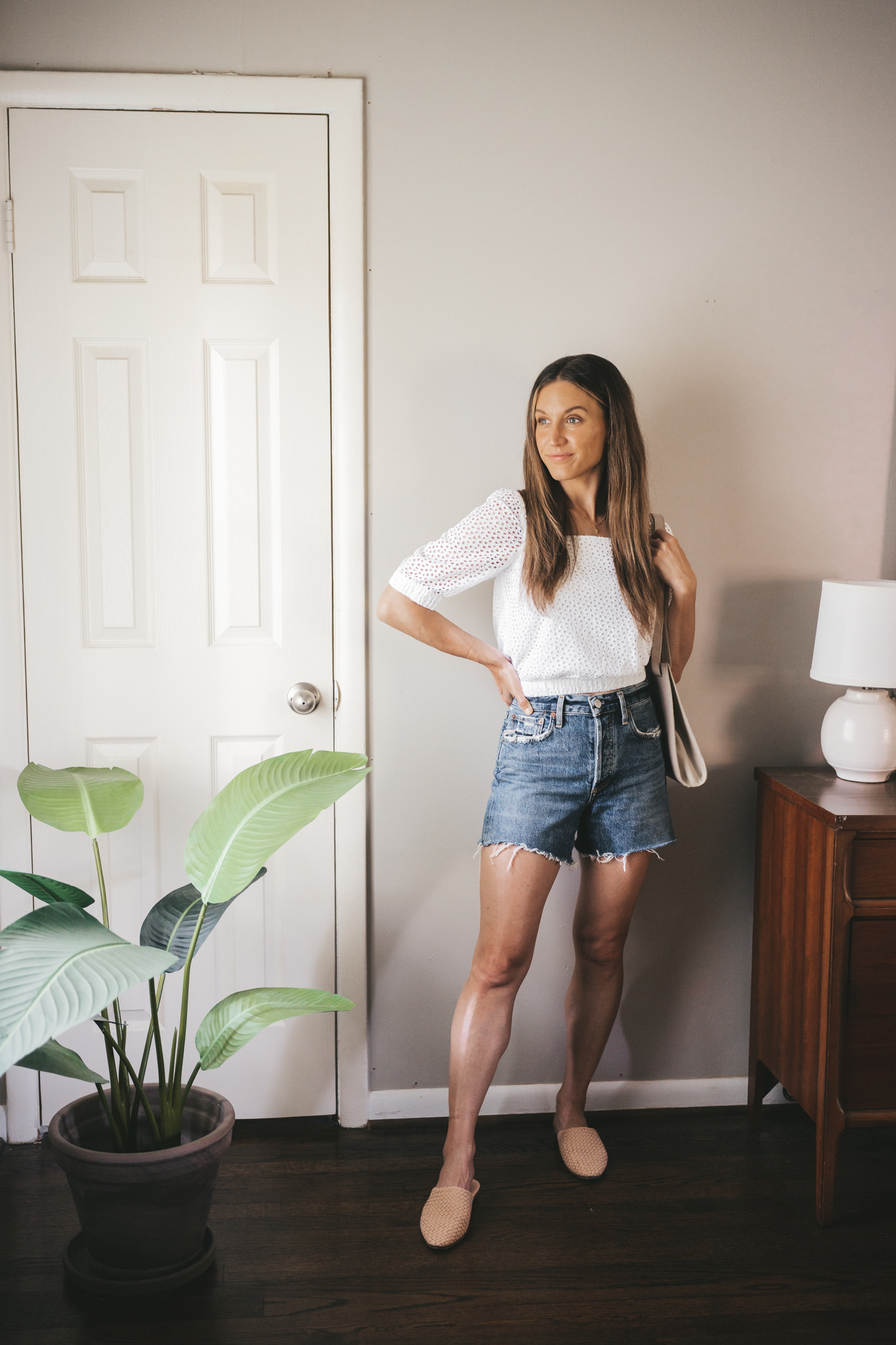 ashley wearing a white puff sleeve eyelet top, mid-wash denim cut-off shorts, woven mules, and a gray shoulder bag