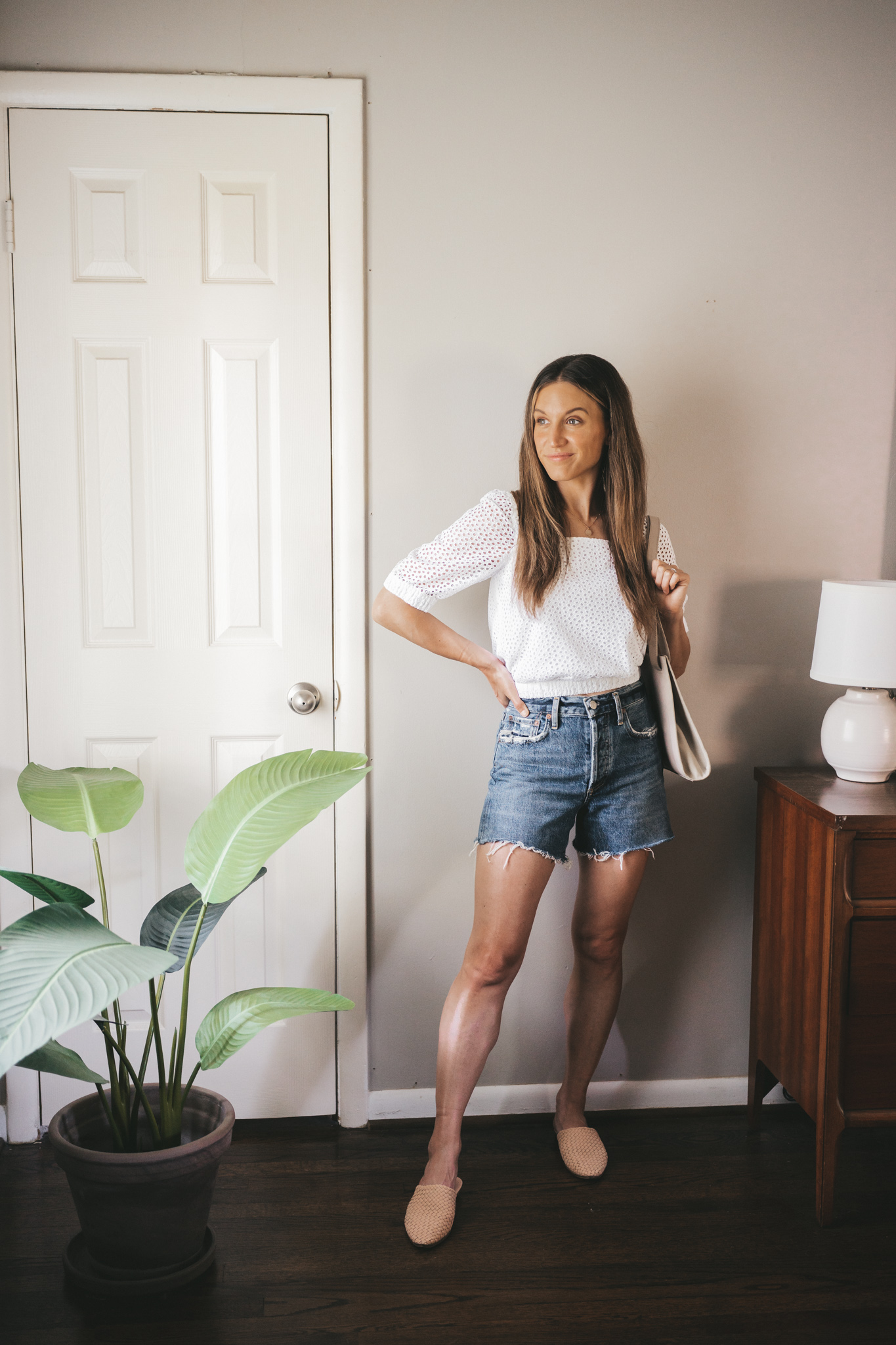 ashley wearing a white puff sleeve eyelet top, mid-wash denim cut-off shorts, woven mules, and a gray shoulder bag