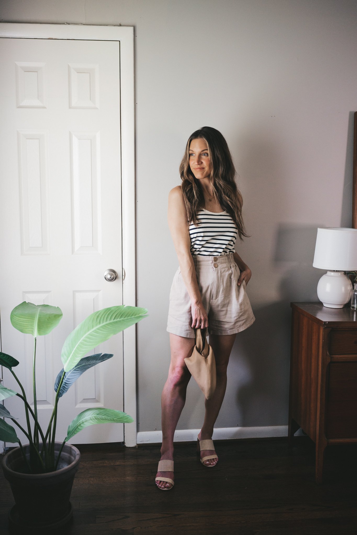 ashley wearing a white and navy striped tank, tan linen shorts, woven heeled sandals, and a tan leather clutch