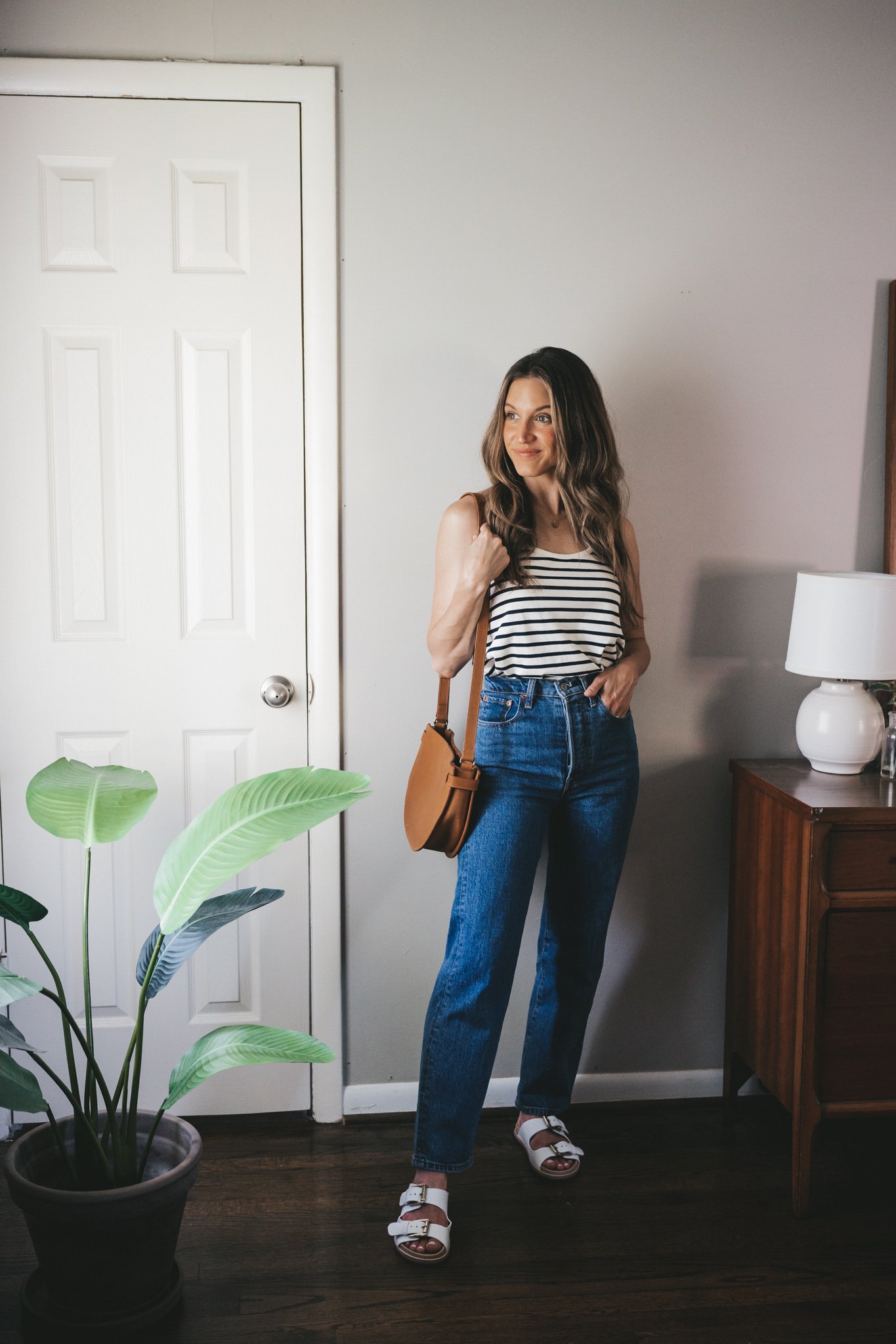 how to wear a striped tank; ashley wearing a white and navy striped tank, mid-wash straight denim, white two strap footbed sandals, and a cognac crossbody bag