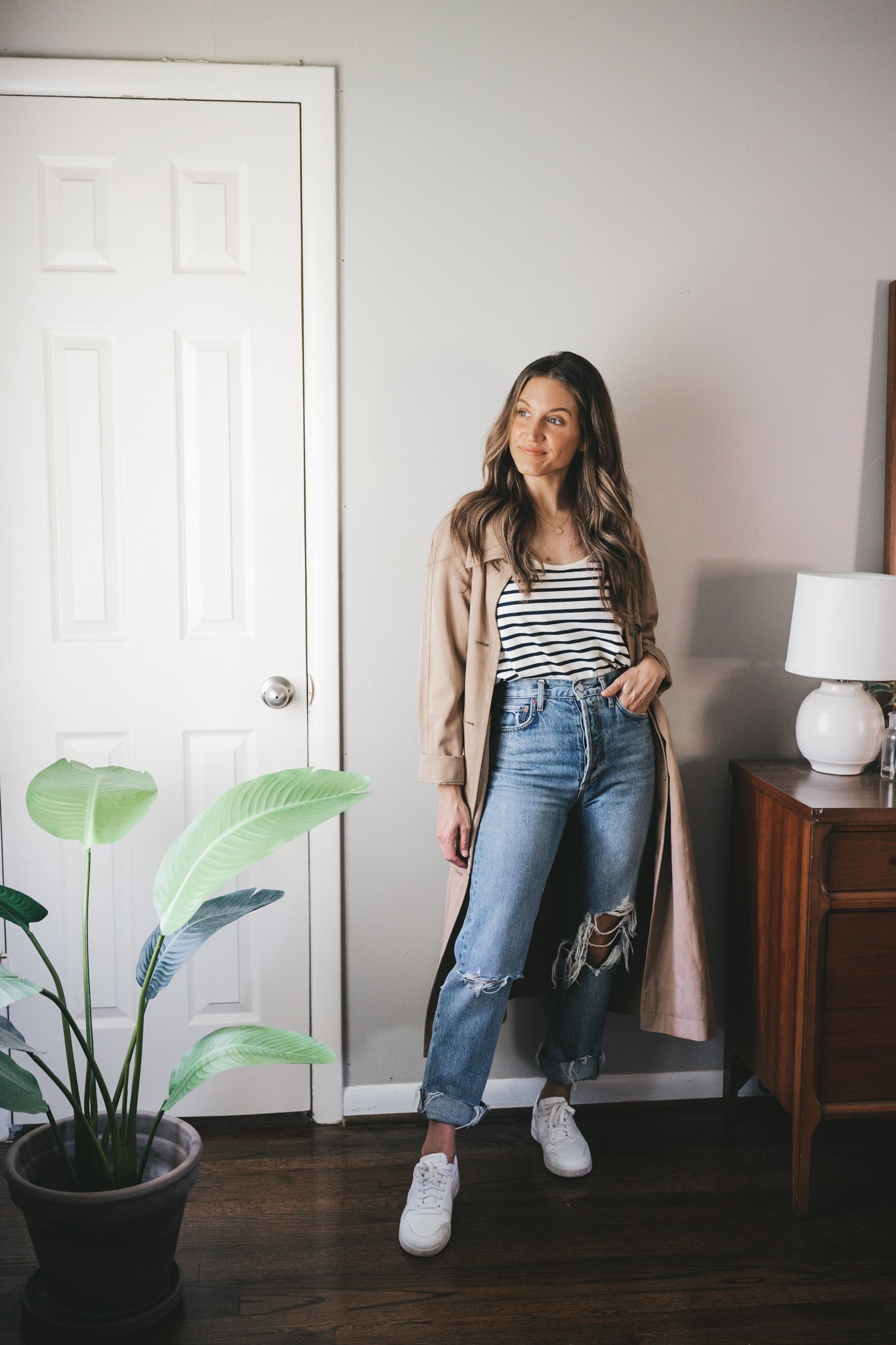 ashley wearing a white and navy striped tank, tan trench coat, distressed denim, and white sneakers