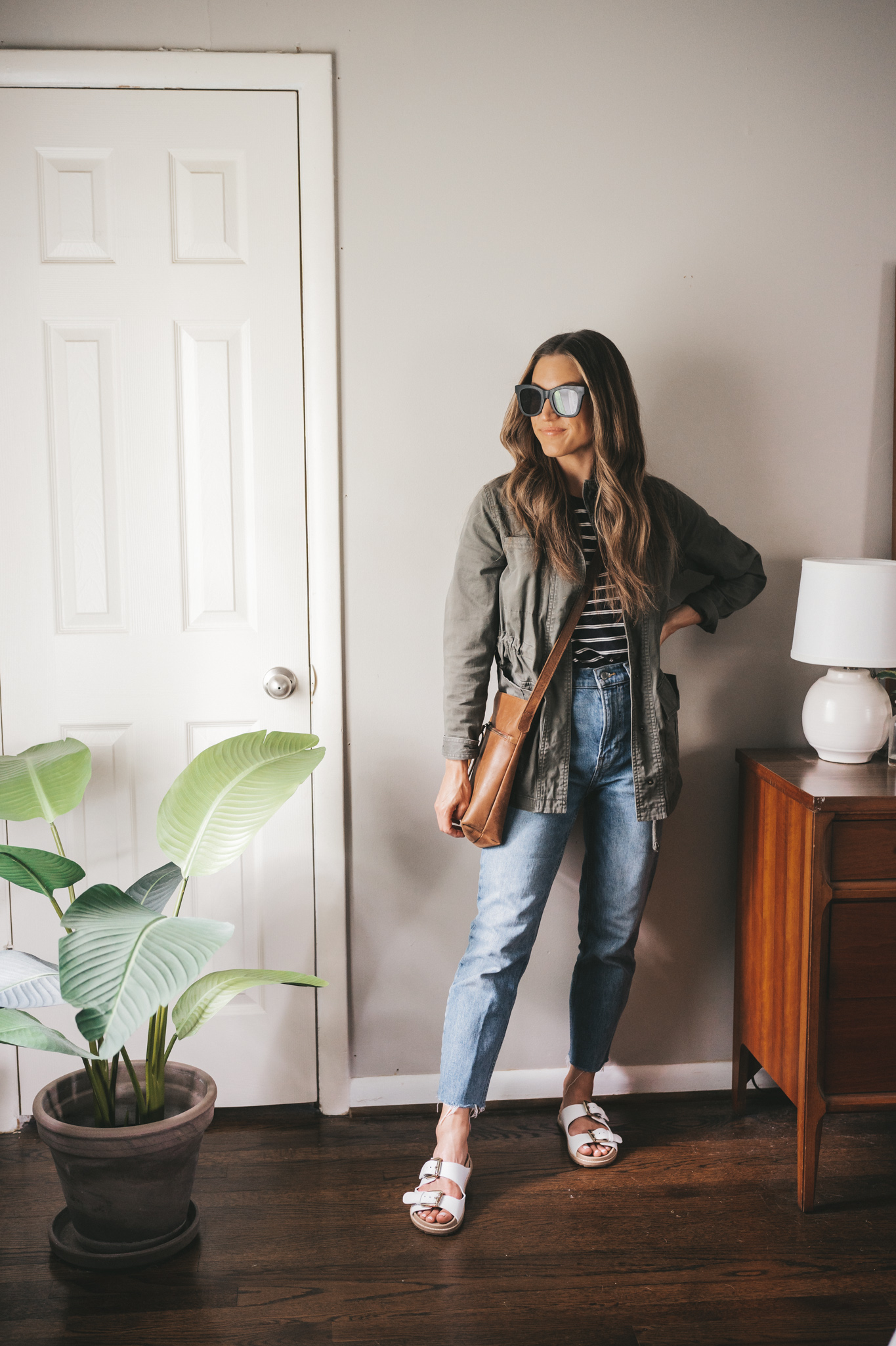 ashley wearing a black and white striped tee, green utility jacket, mid-wash straight denim, and white sandals with a brown leather crossbody bag
