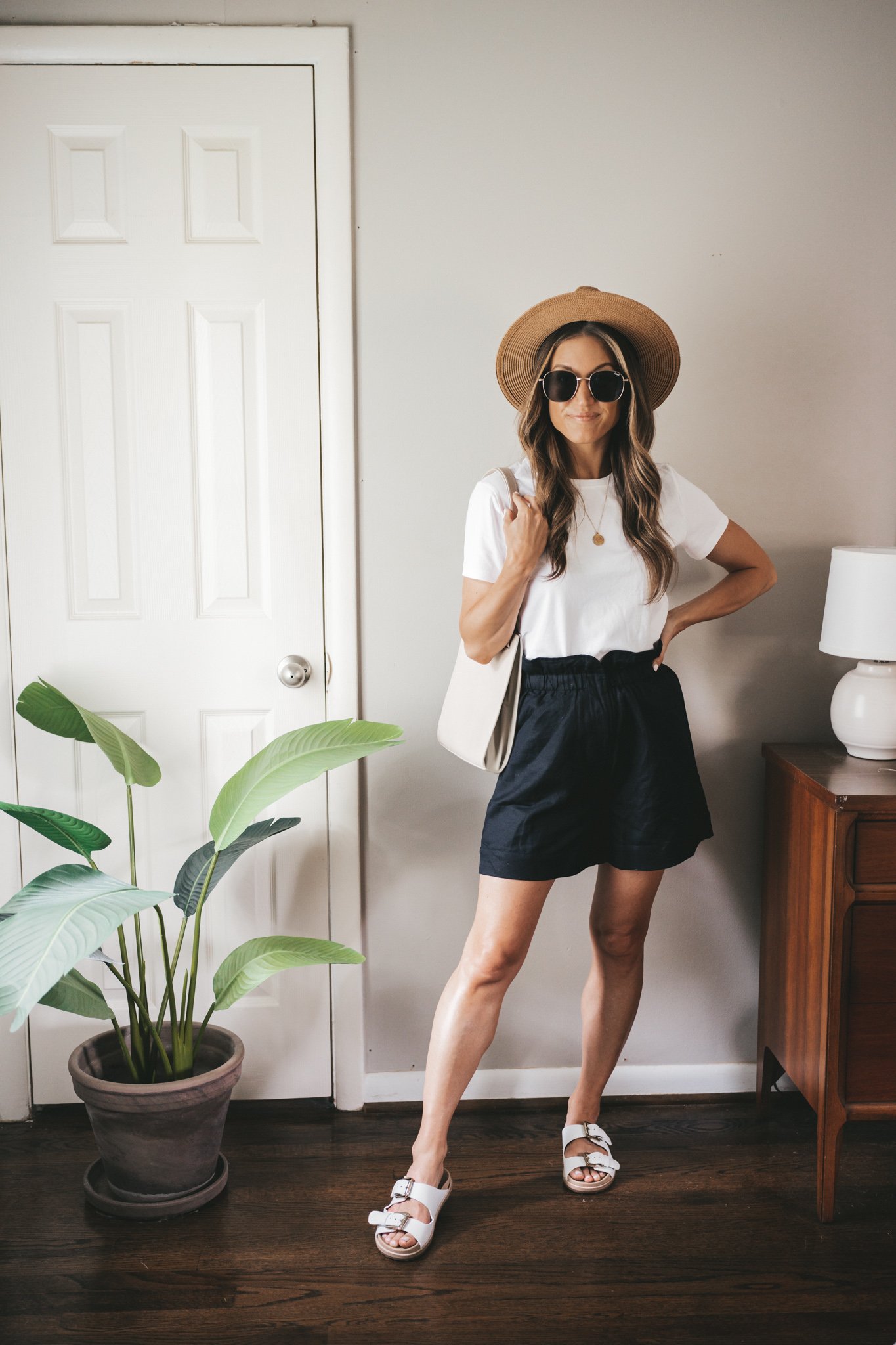 ashley wearing a white tee, black paperbag shorts, white sandals, and a gray crossbody bag with a straw fedora hat