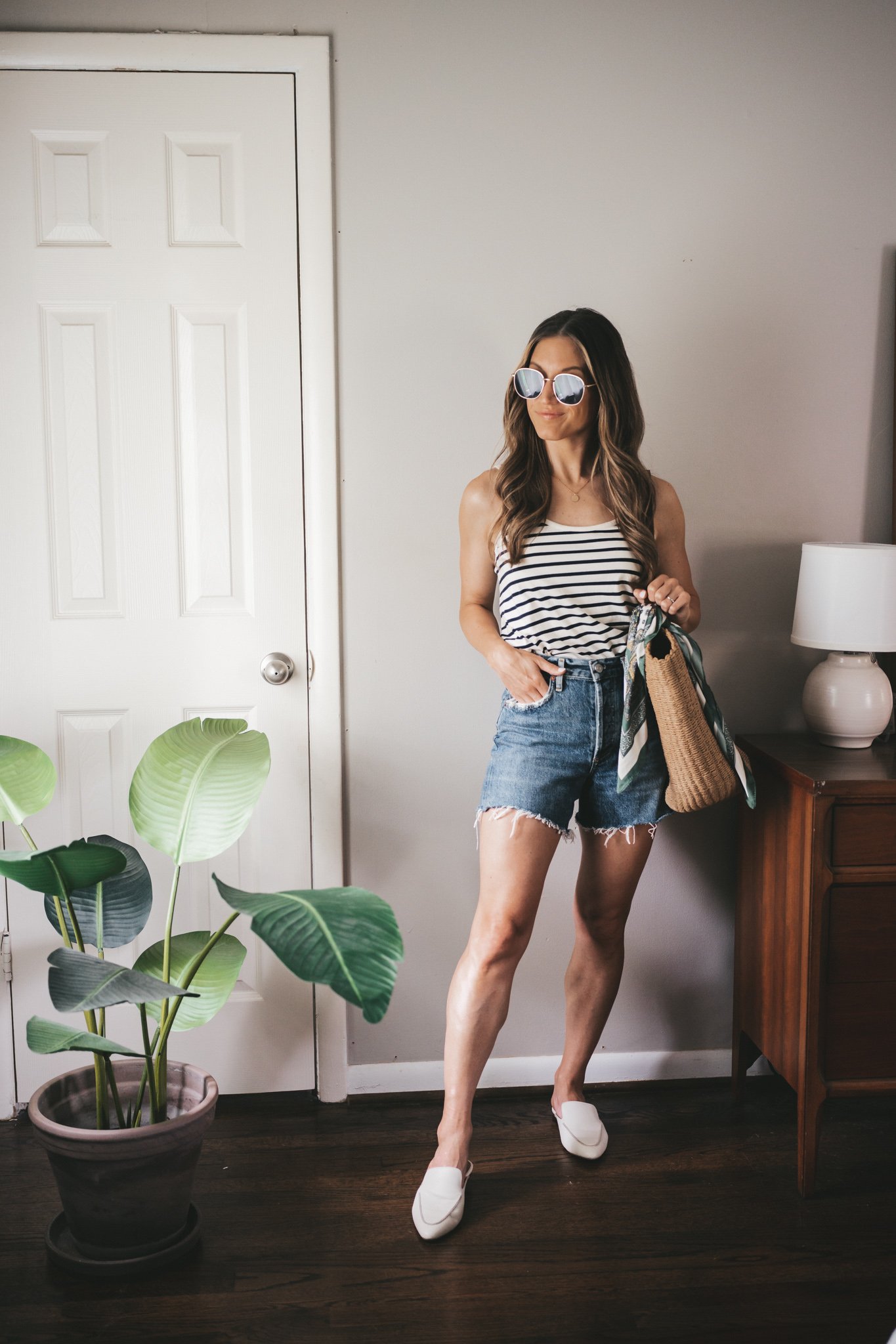 ashley wearing a white and navy striped tank, denim cut-off shorts, white mules, a straw tote bag with a scarf tied around the handle, and oversized gold framed sunglasses