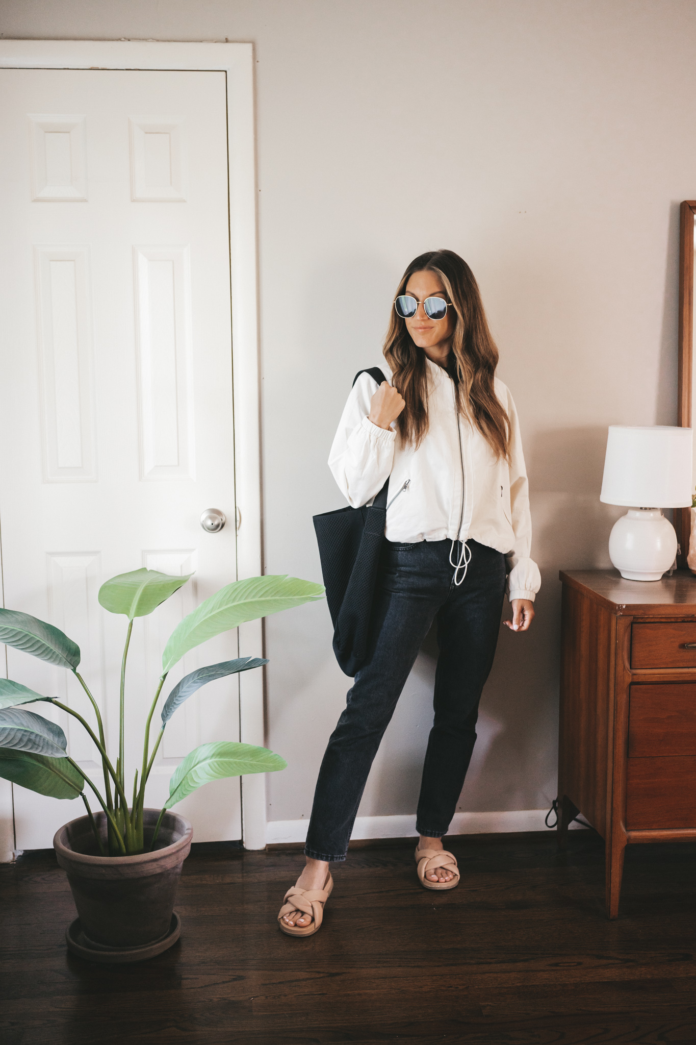 ashley wearing a white bomber jacket, washed black denim, and padded slides with a black tote bag