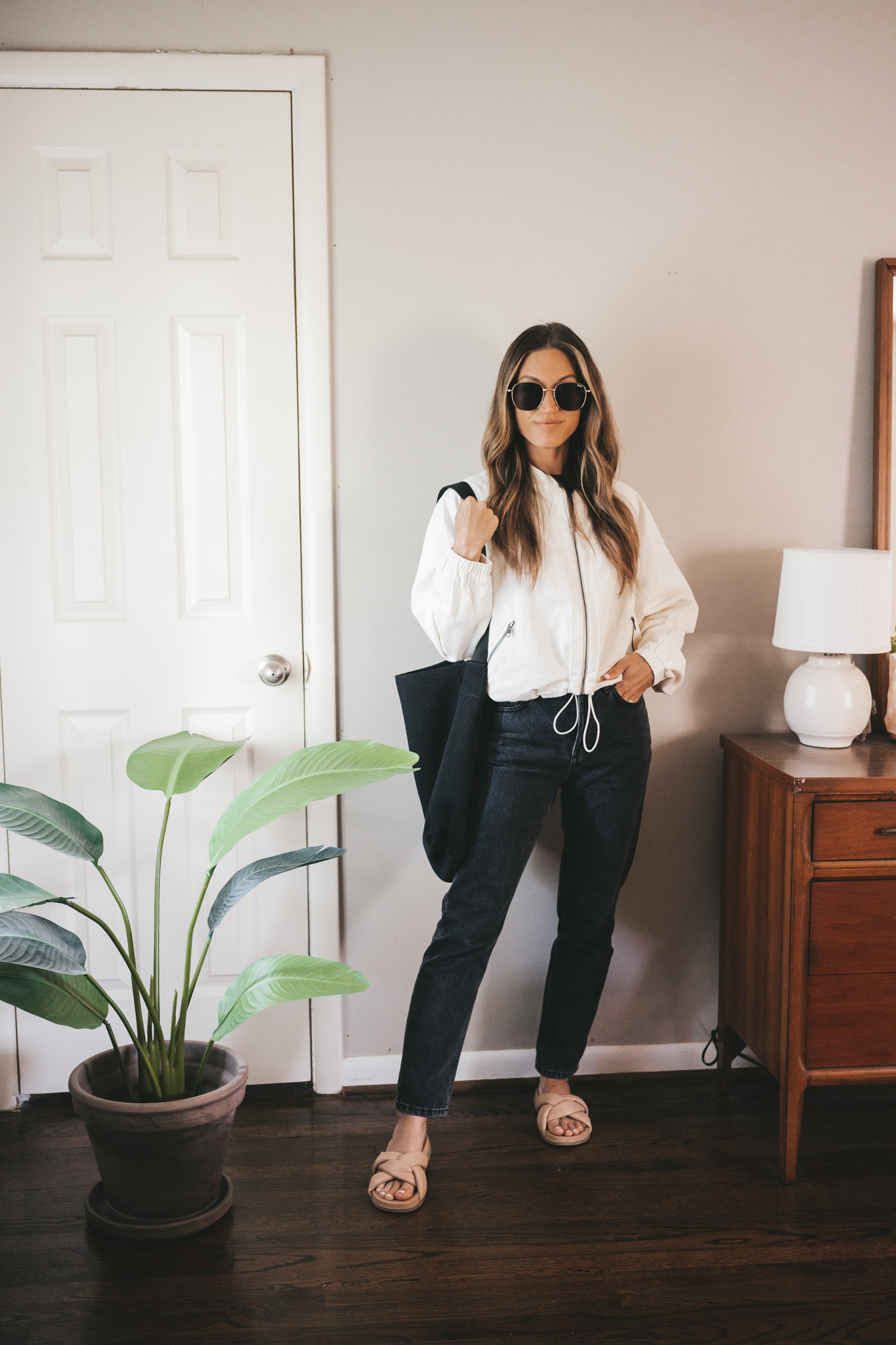 ashley wearing a white bomber jacket, washed black denim, and padded slides with a black tote bag