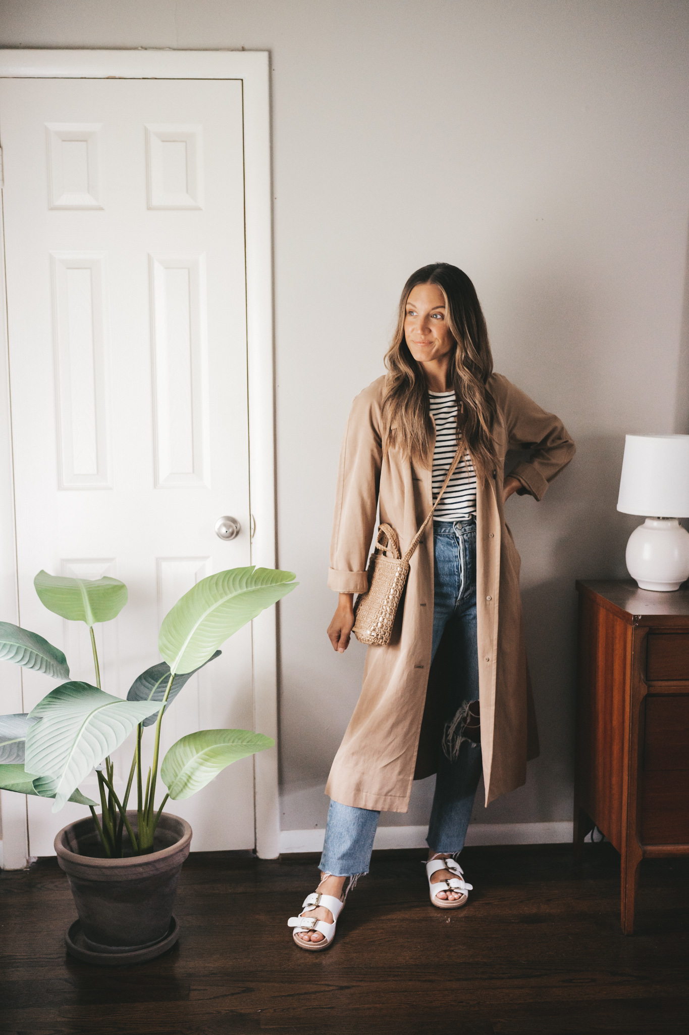 ashley wearing a striped tee, trench coat, distressed denim, and white two strap sandals with a woven crossbody bag