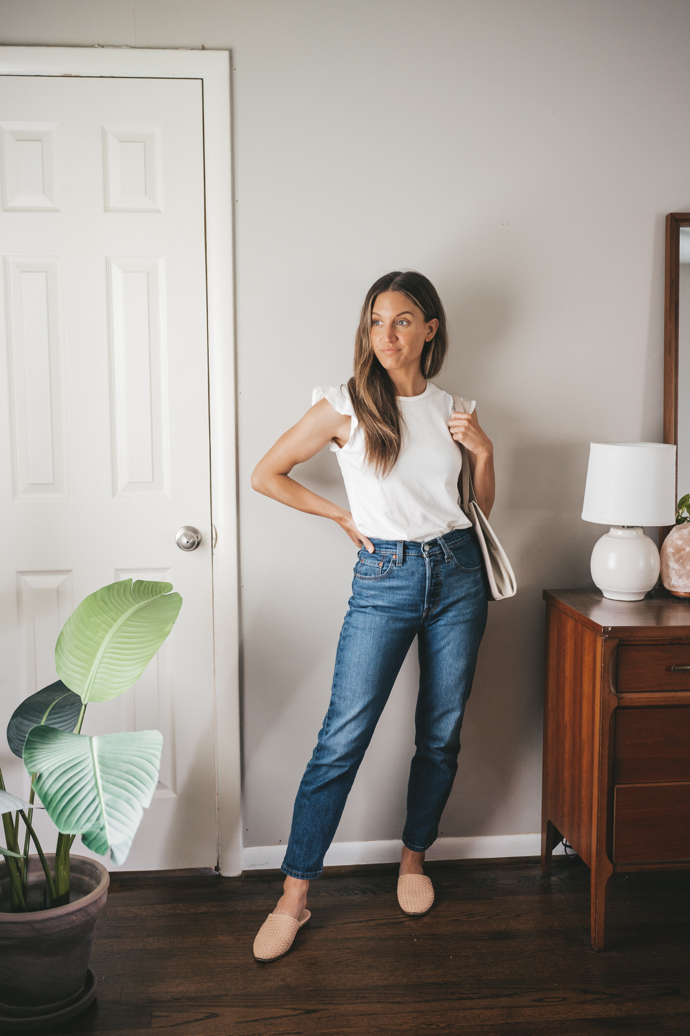 ashley wearing a white tank with ruffles on the shoulders, dark wash straight denim, woven mules, and a gray shoulder bag