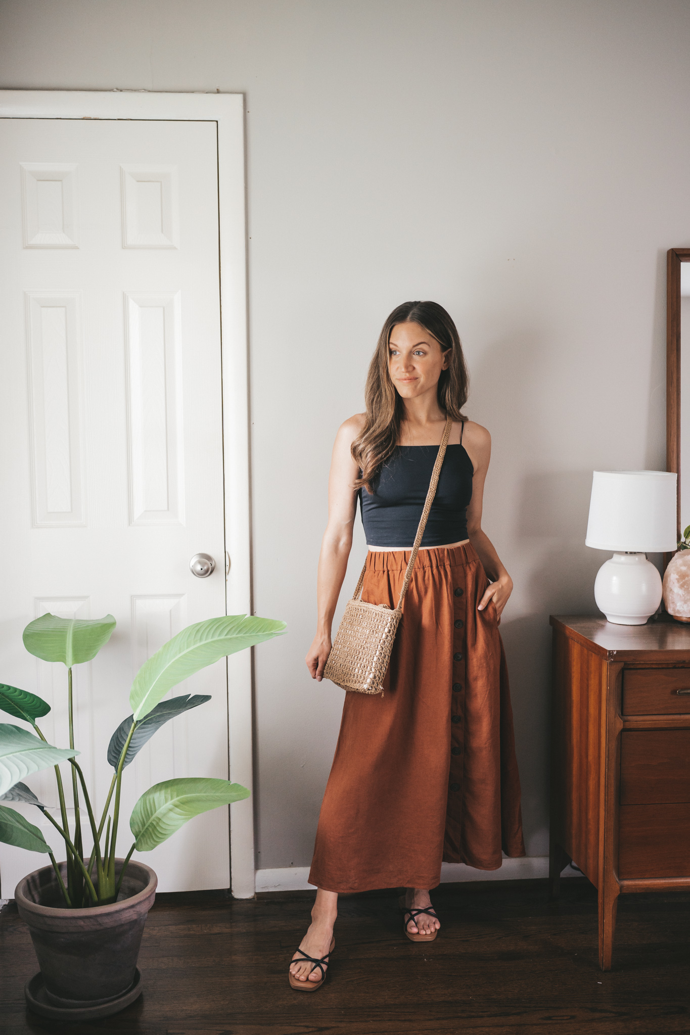 ashley wearing a black cropped cami, rust colored midi skirt, black and tan sandals, and a woven crossbody bag