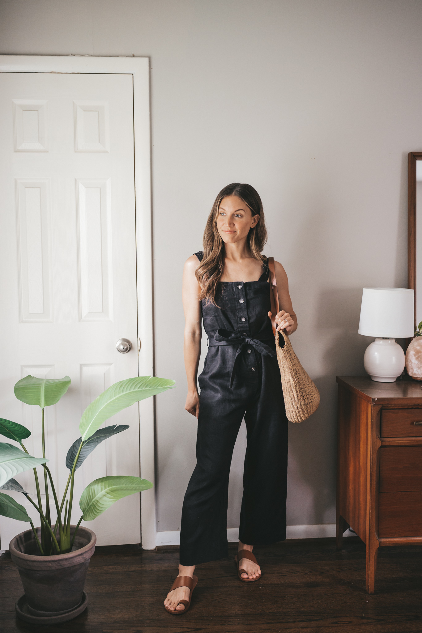 ashley wearing a black jumpsuit, brown leather sandals, and a straw tote bag