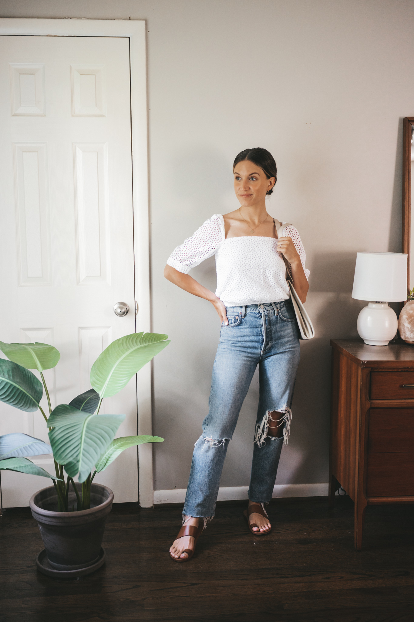 ashley wearing an puff sleeve eyelet top, distressed denim, and brown leather sandals with a gray shoulder bag