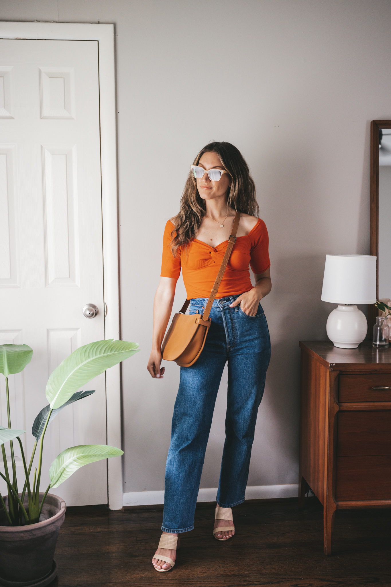 ashley wearing a coral red top, mid-wash denim, woven heeled sandals, and a cognac crossbody bag