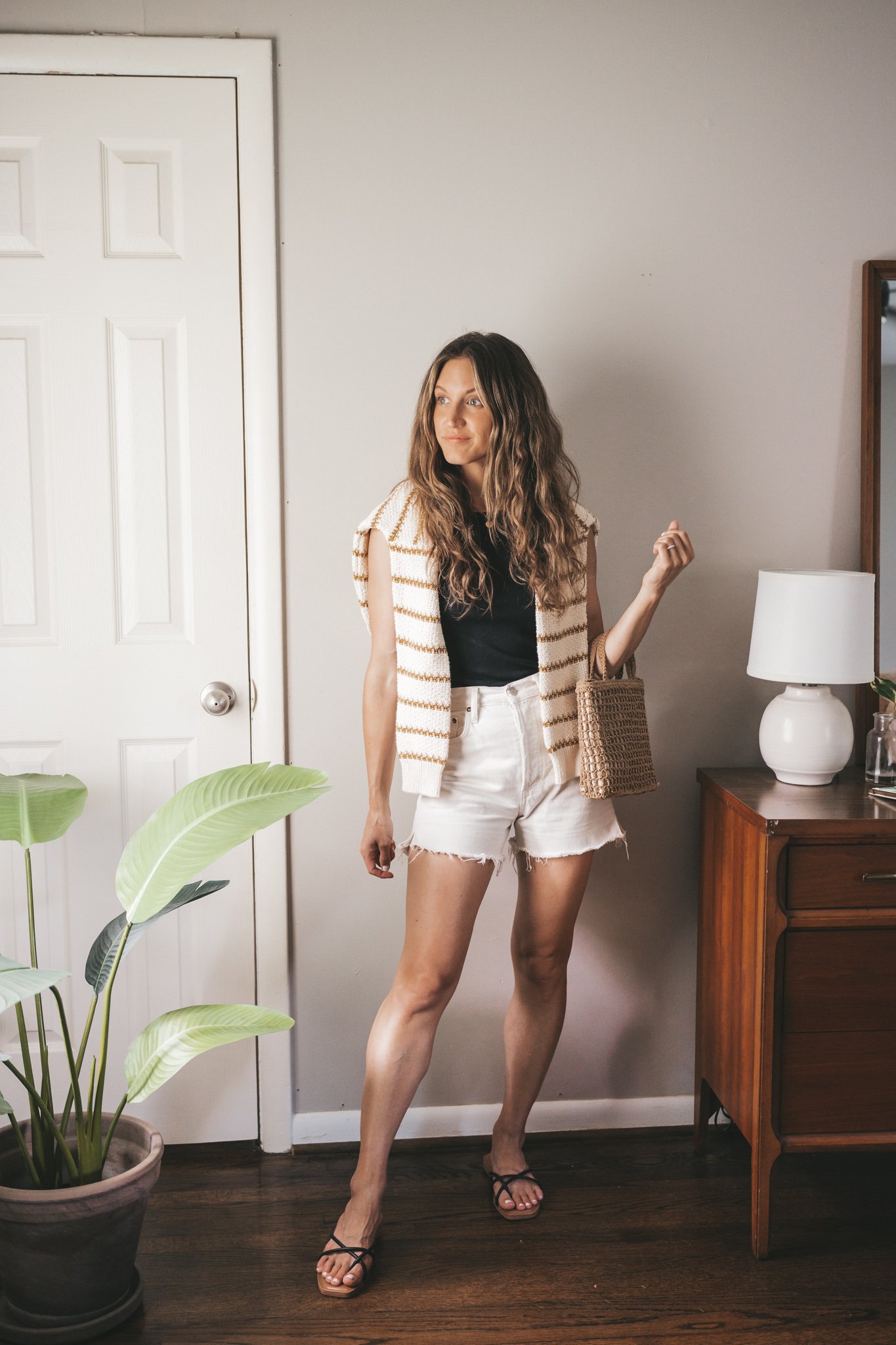 ashley wearing a black tee, white denim shorts, strappy black sandals, a striped sweater worn over the shoulders, and a straw handbag