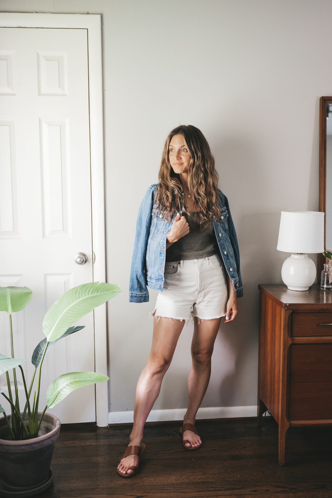ashley wearing a taupe sweater tank, denim jacket, white denim shorts, and brown leather sandals