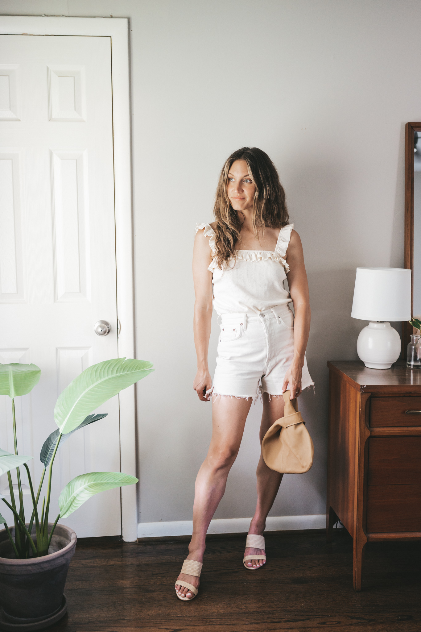 ashley wearing a white tank with ruffle straps, white denim shorts, woven heeled sandals, and a leather clutch
