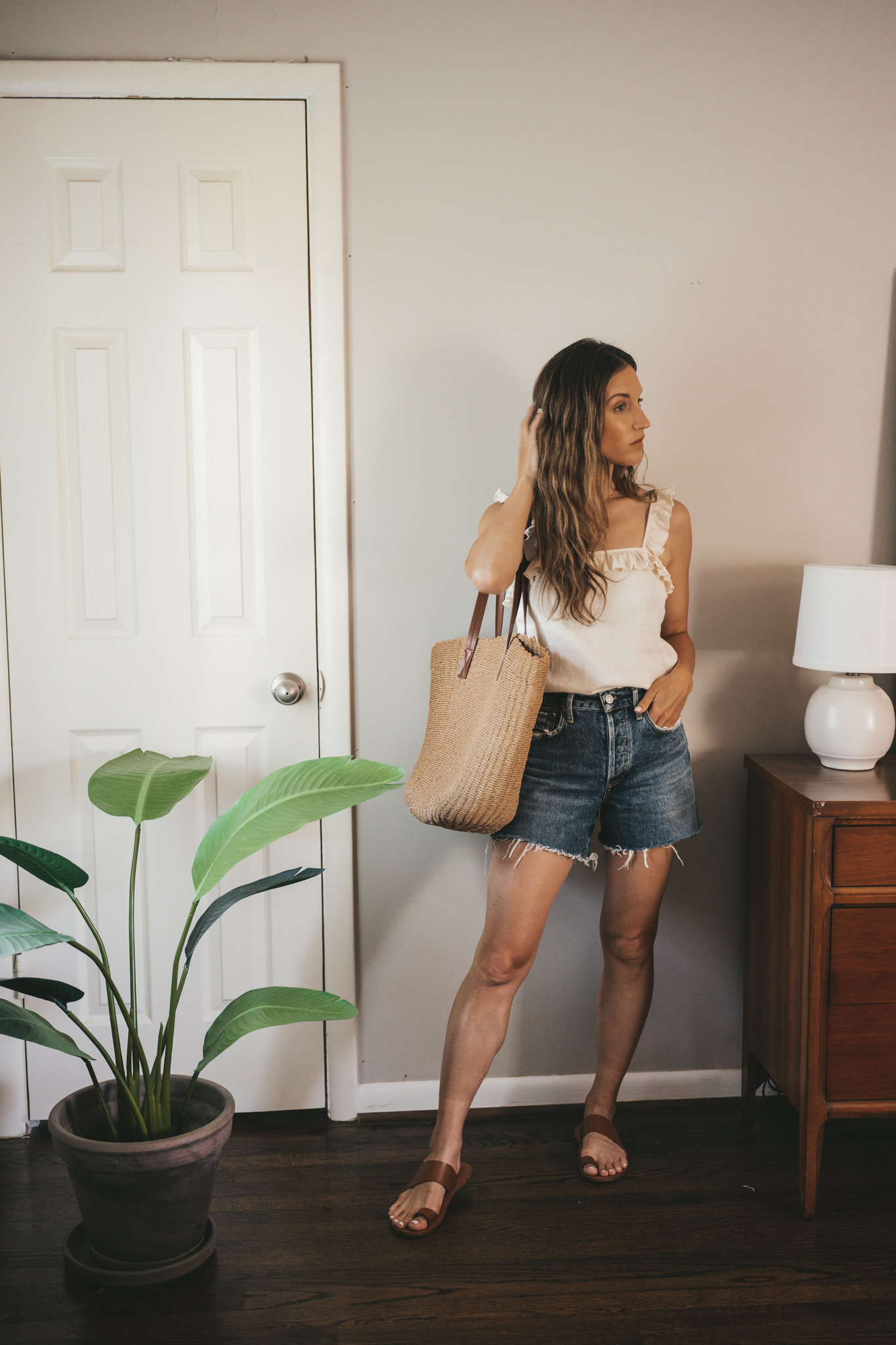 ashley wearing a white tank with ruffle straps, denim shorts, brown leather sandals, and a straw tote bag