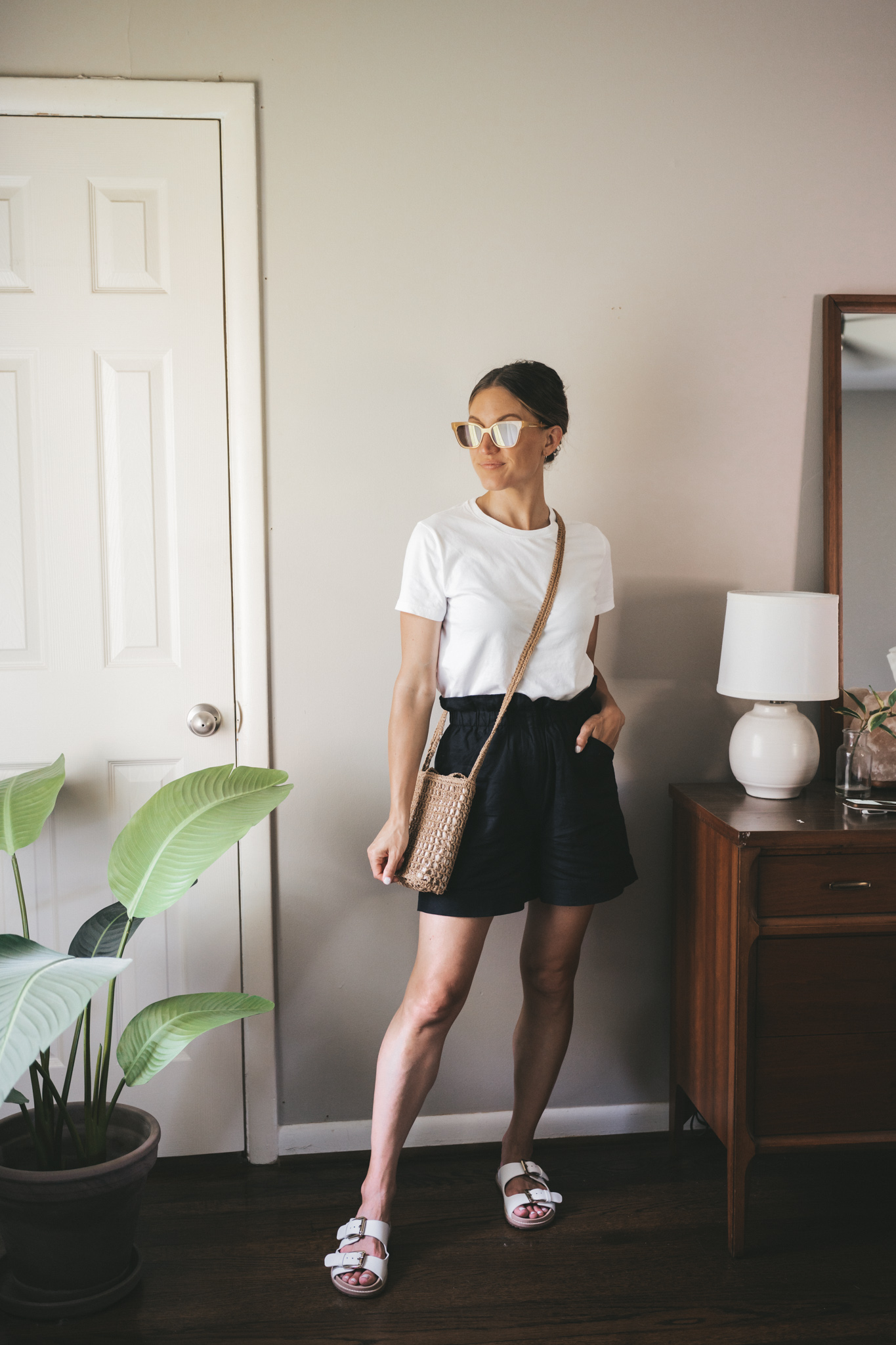ashley wearing a white tee, black paperbag shorts, white sandals, and a straw crossbody bag