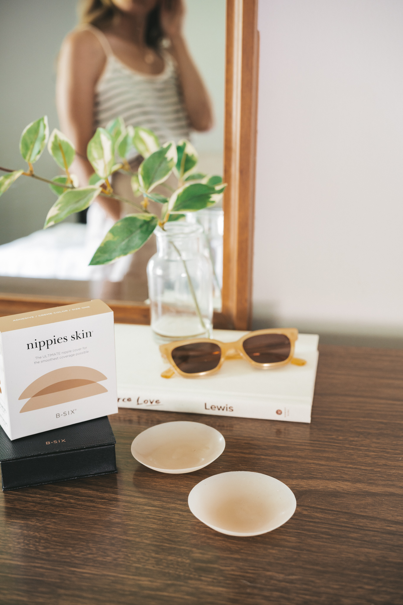 a dresser with a mirror, and a package of Nippies Nipple Covers, a white book, a pair of sunglasses, and a vase with a faux plant stem