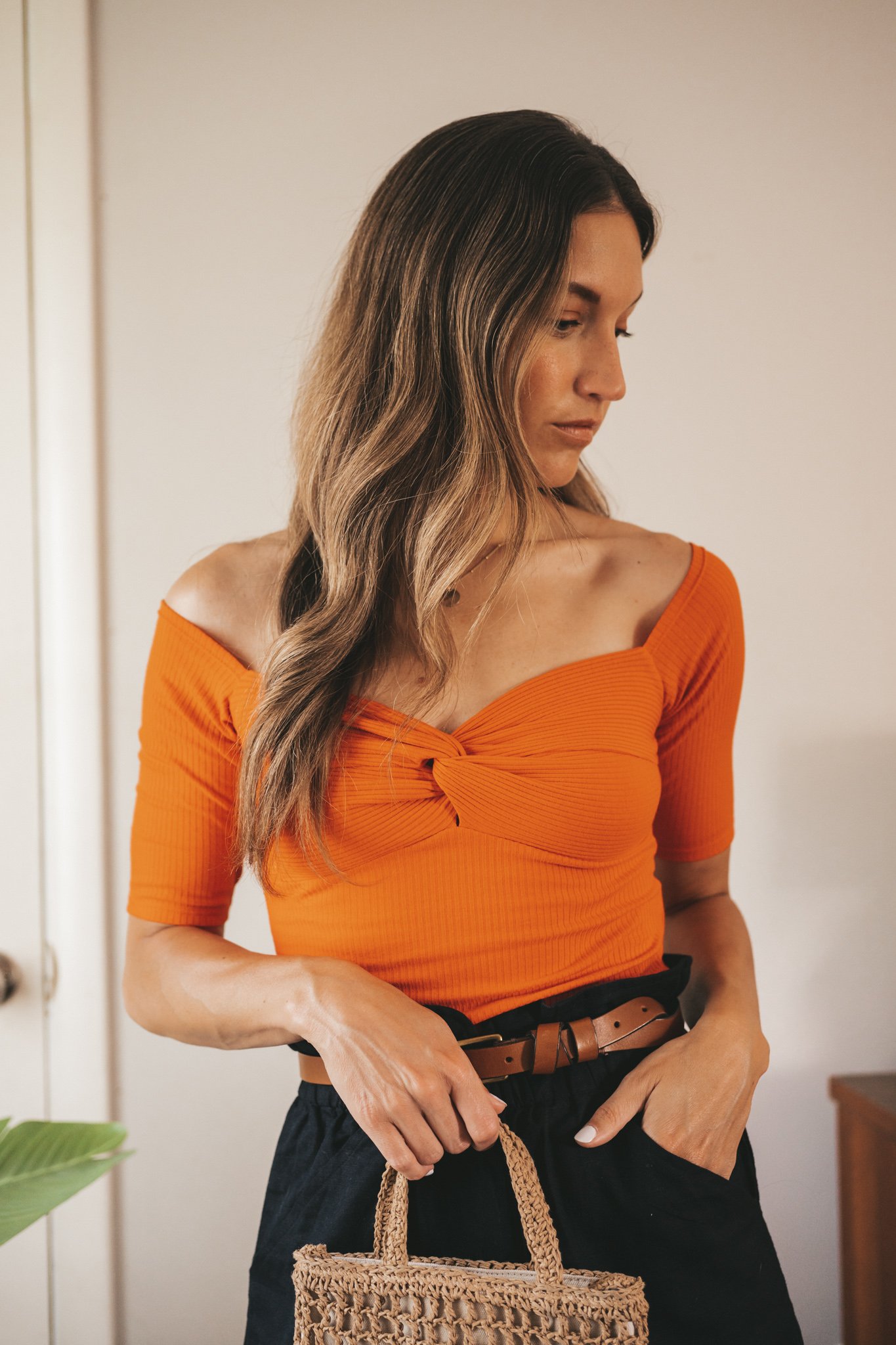 ashley wearing a red top with a sweetheart neckline, black paperbag shorts, a brown belt, and a woven handbag
