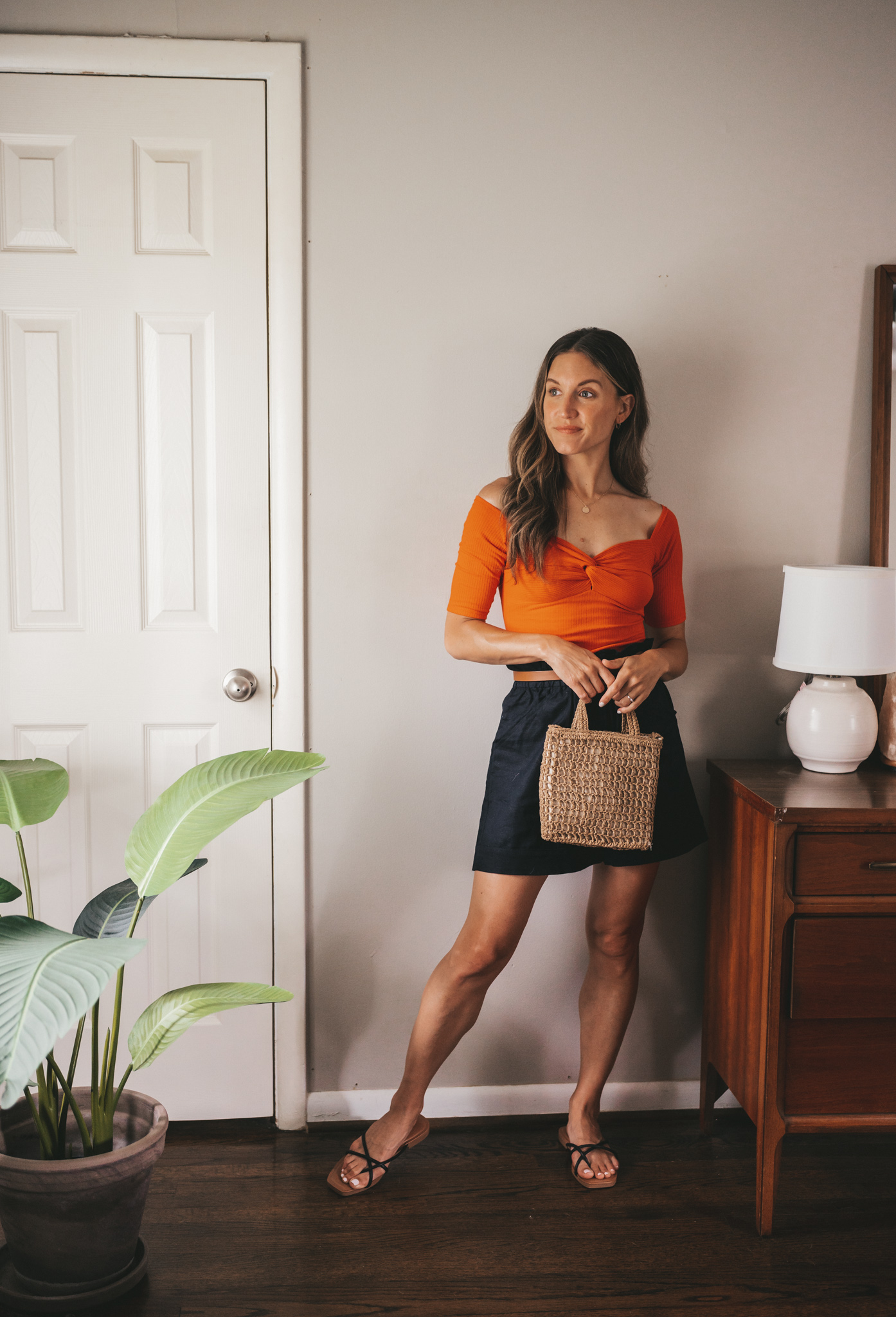 ashley wearing a red top with a sweetheart neckline, black paperbag shorts, a brown belt, and a woven handbag